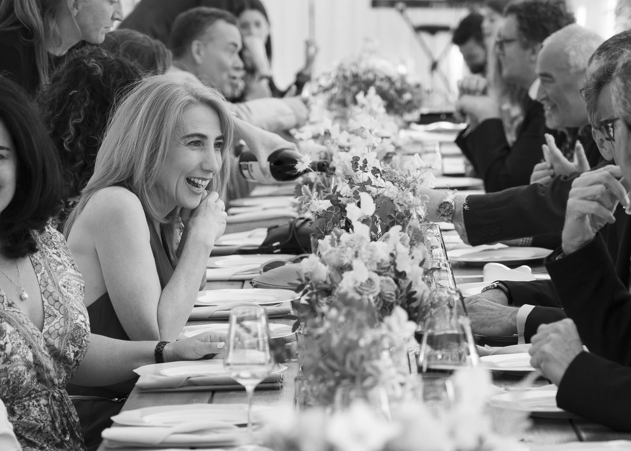 A group of people sitting at a long table decorated with flowers, enjoying a meal and drinks at a social gathering.
