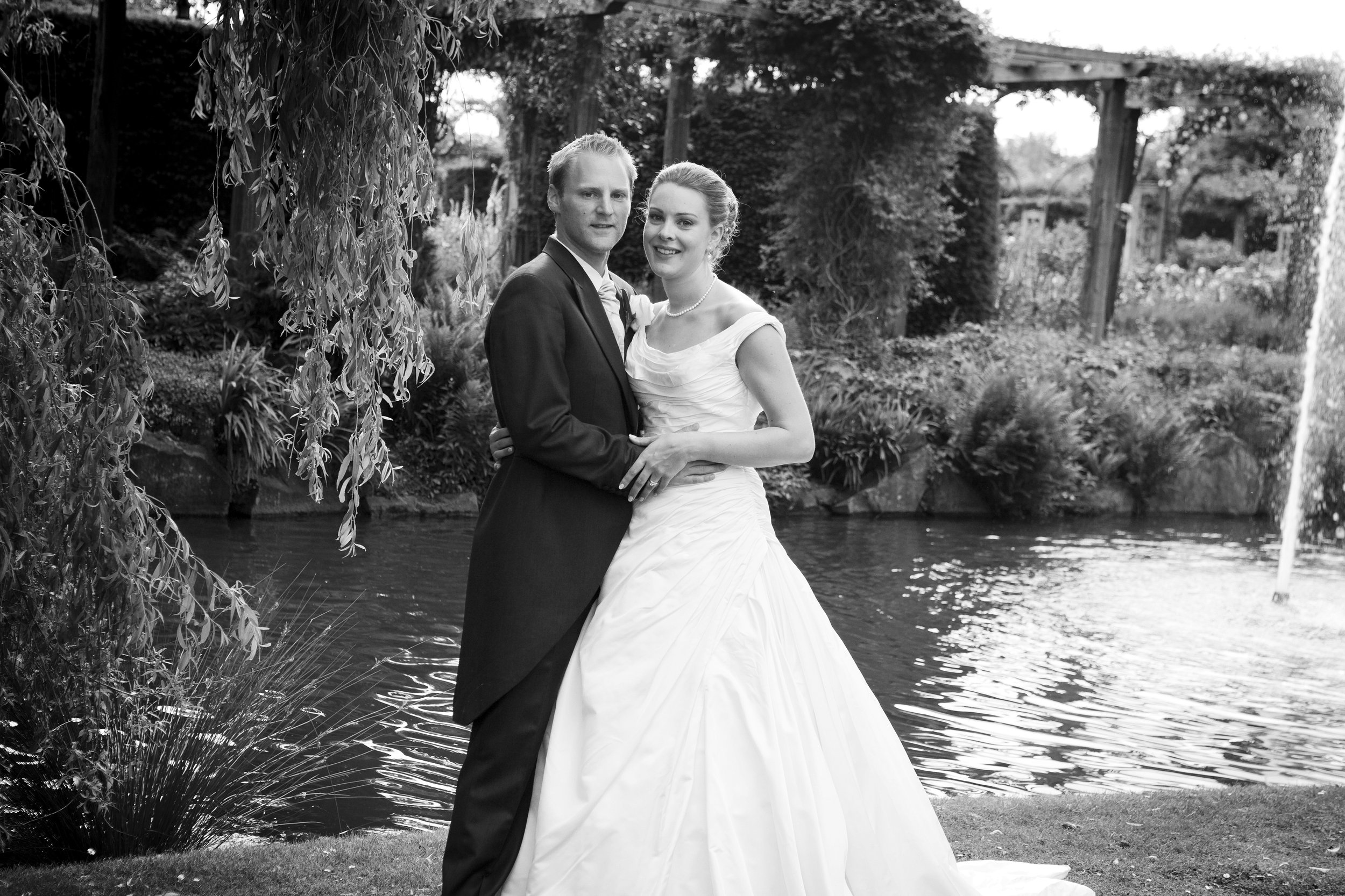 A black and white photo of a bride and groom standing by a pond in a garden. The bride wears a white wedding gown and the groom is in a dark suit. They are smiling and holding hands.