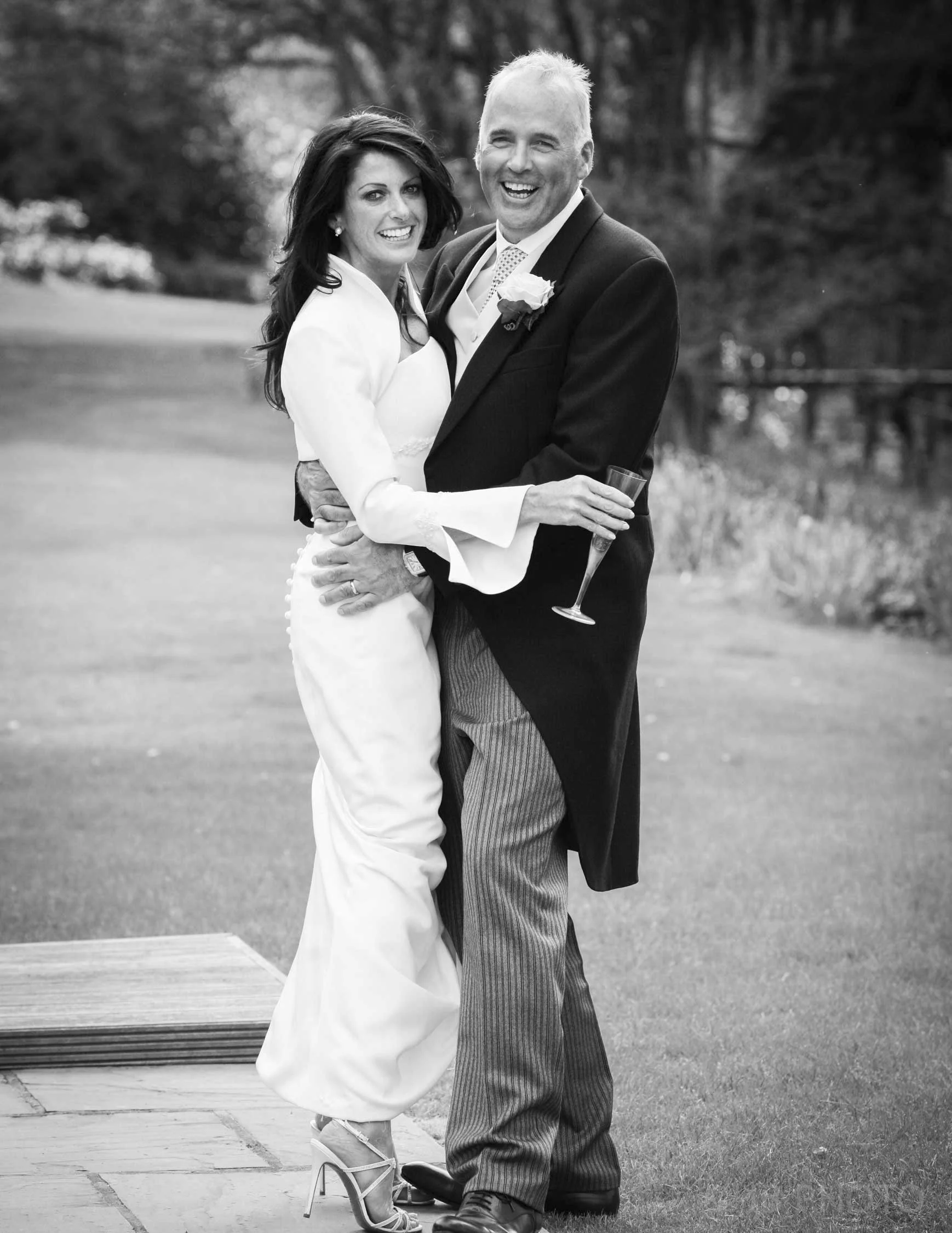 Black and white photo of a smiling bride and groom standing outdoors, with the bride holding a champagne flute and the groom wearing a boutonniere, surrounded by trees.