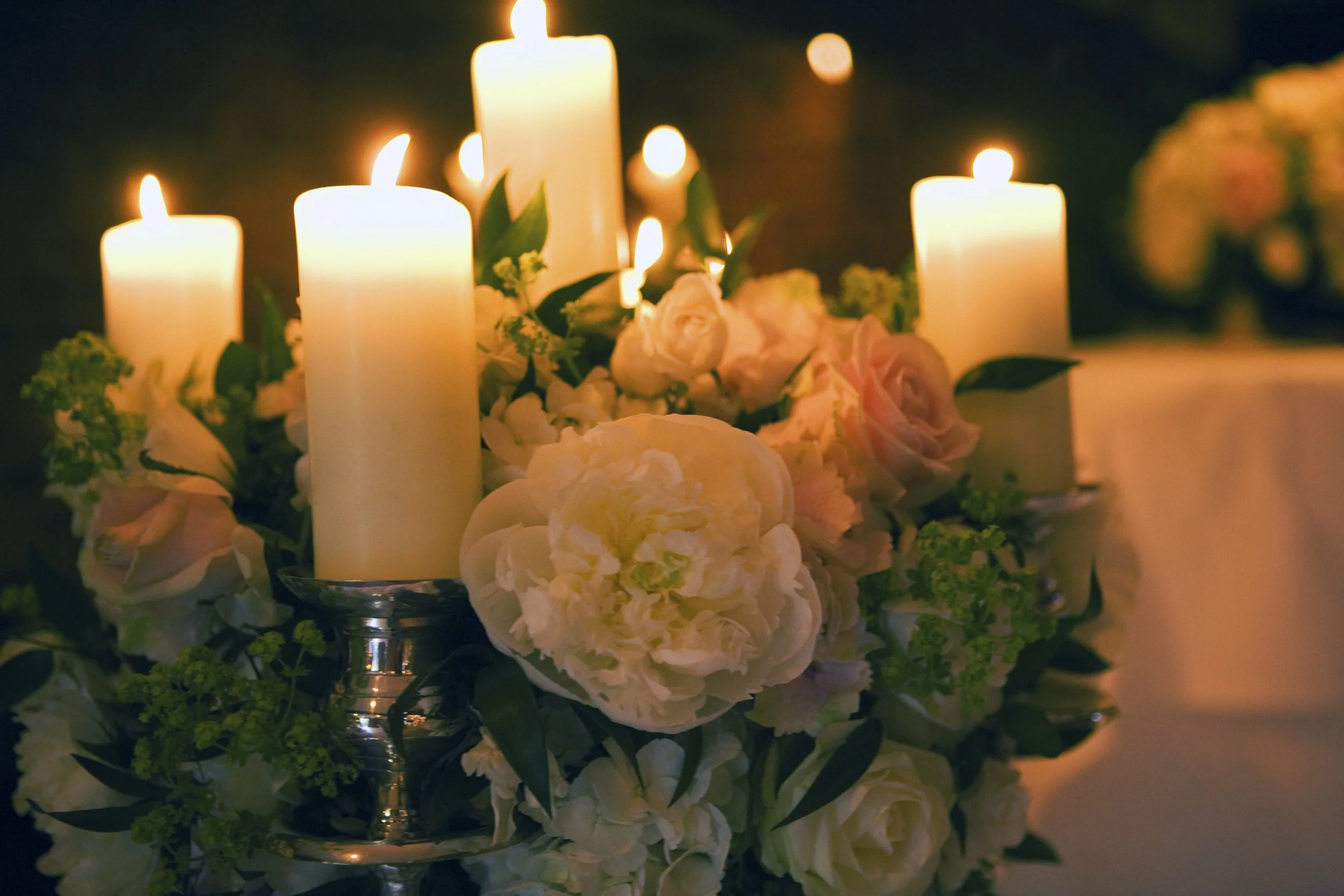 A floral centerpiece with white and pale pink flowers, including roses and peonies, illuminated by four lit white candles in silver holders at a dimly lit event or gathering.