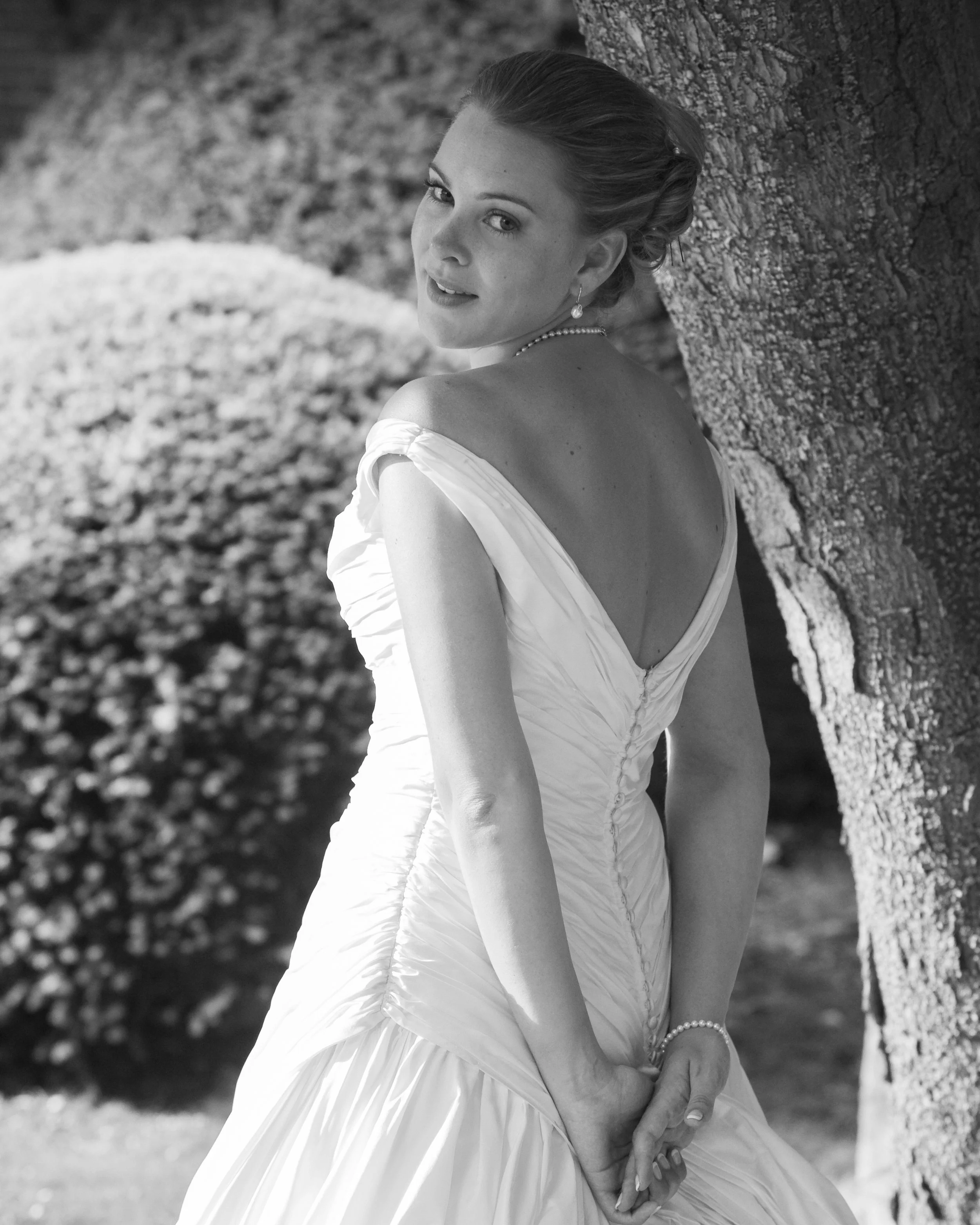 A woman in a wedding dress standing outdoors near a tree, smiling at the camera, with bushes in the background.