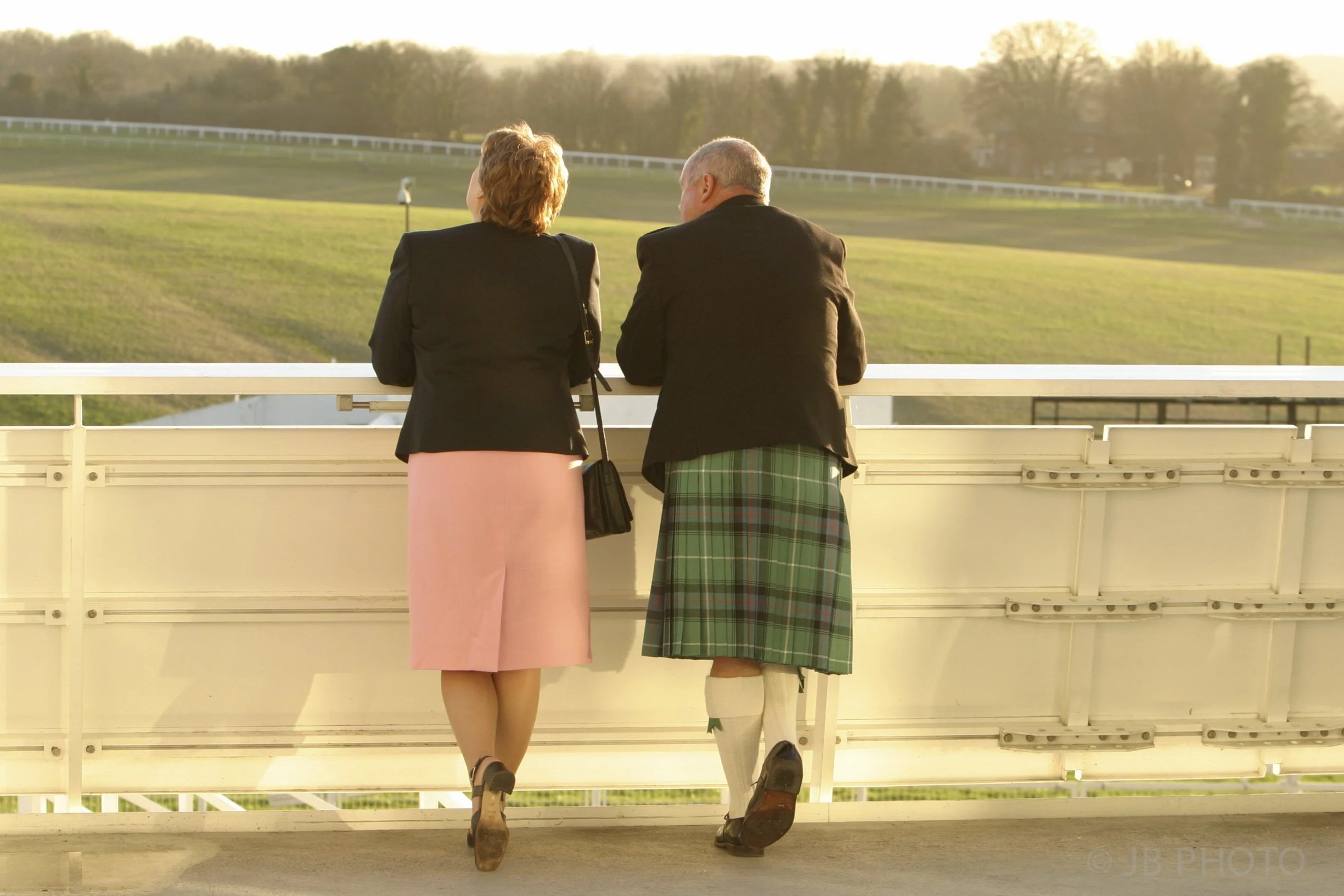 A woman and man standing on a balcony overlooking a grassy racetrack, with the woman wearing a black blazer, pink skirt, and heels, and the man in a black blazer, kilt, and socks, during the late afternoon or early evening.
