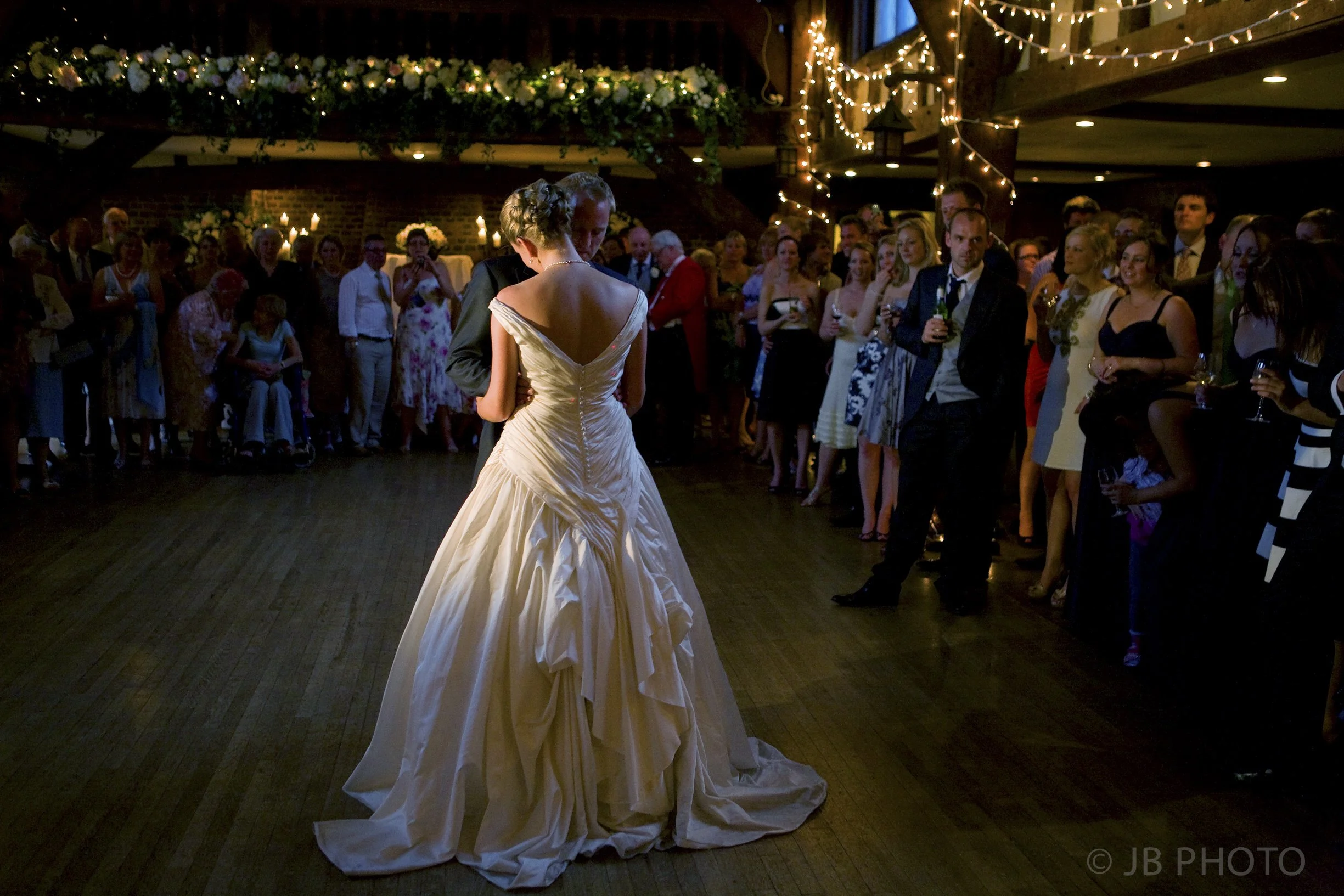 A bride and groom sharing their first dance at a wedding reception, surrounded by guests watching in a decorated venue with string lights and floral arrangements.