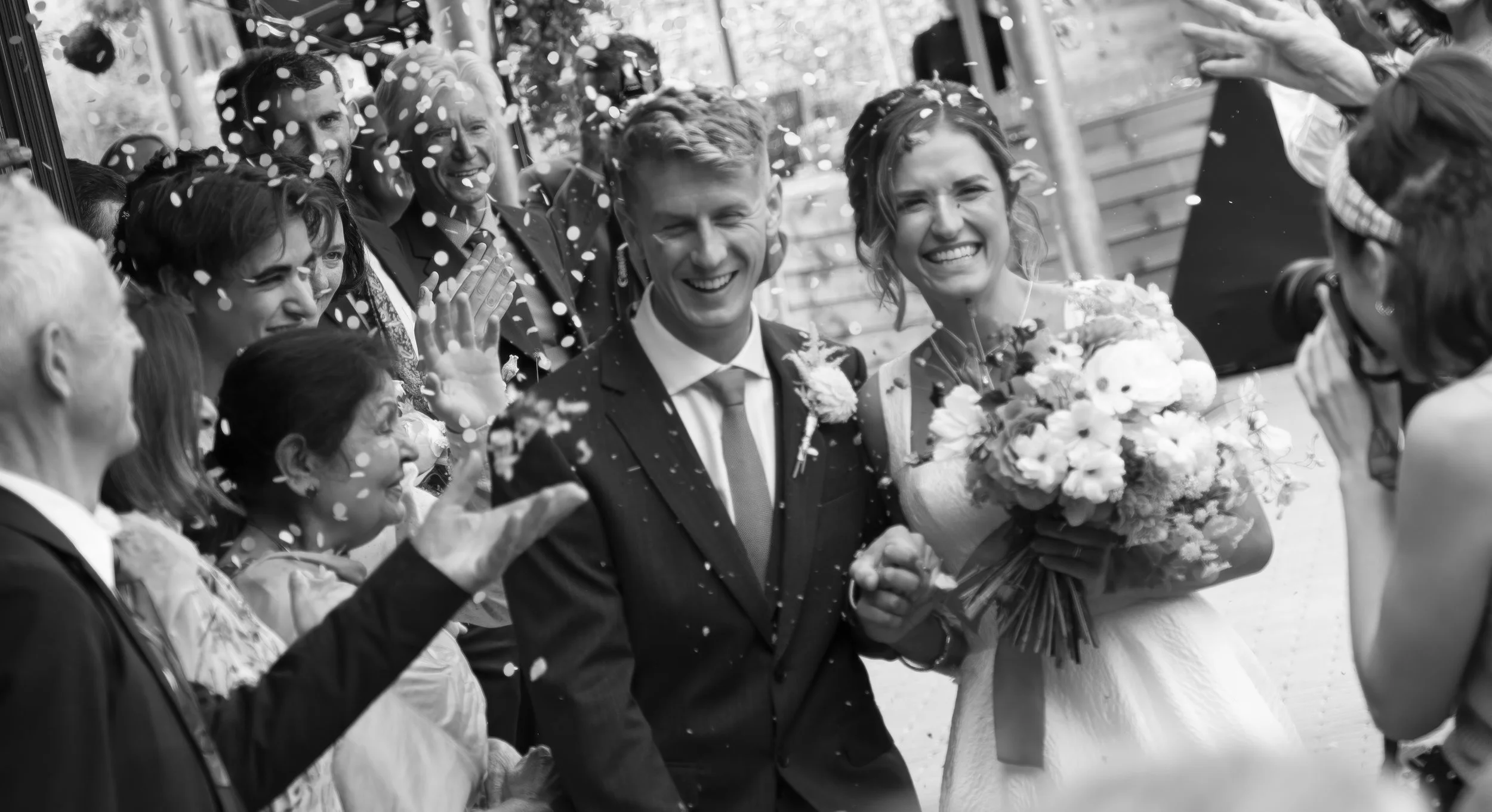 Black and white photo of a bride and groom smiling as they walk through a crowd of wedding guests, confetti falling around them, the bride holding a bouquet of flowers.