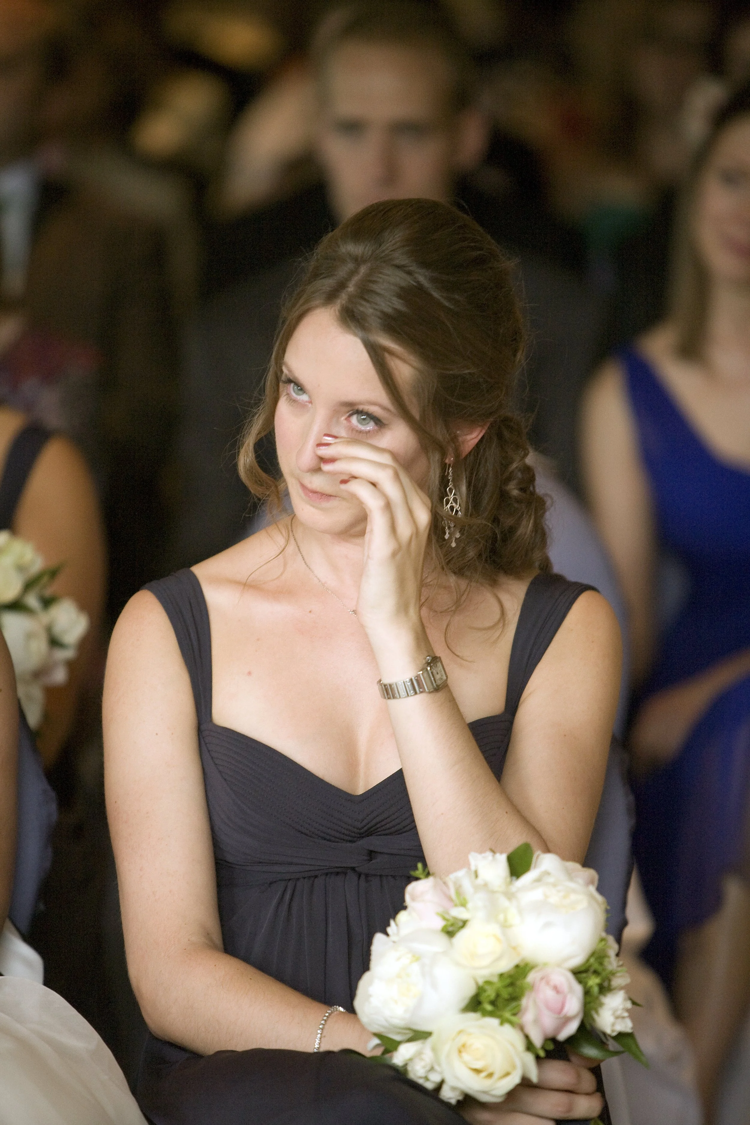 A woman with brown hair in a black dress sitting at a formal event, possibly a wedding, wiping tears from her eyes while holding a bouquets of white and pink flowers, with other guests in the background.