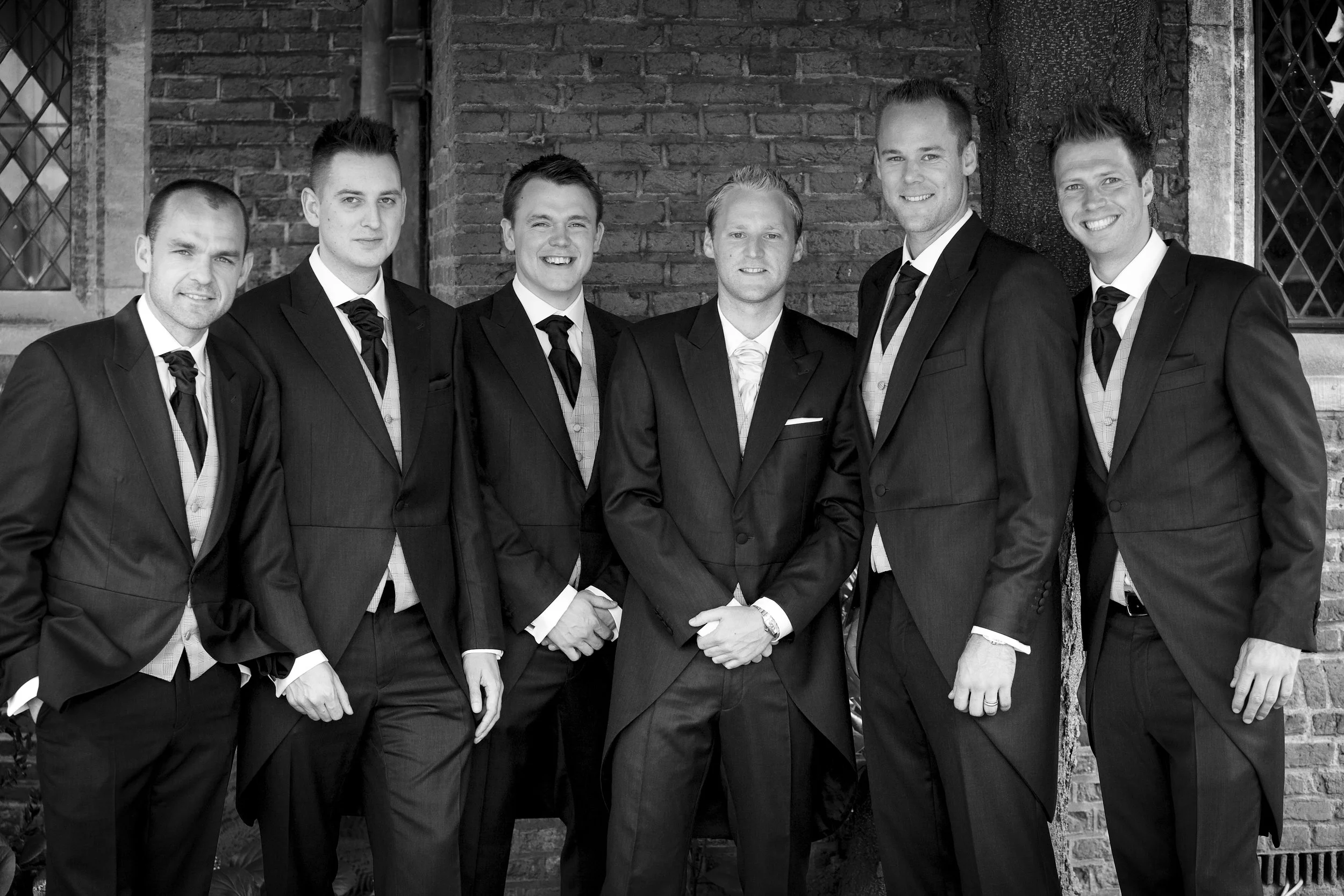 Seven men dressed in formal suits posing outside in front of a brick wall and window.