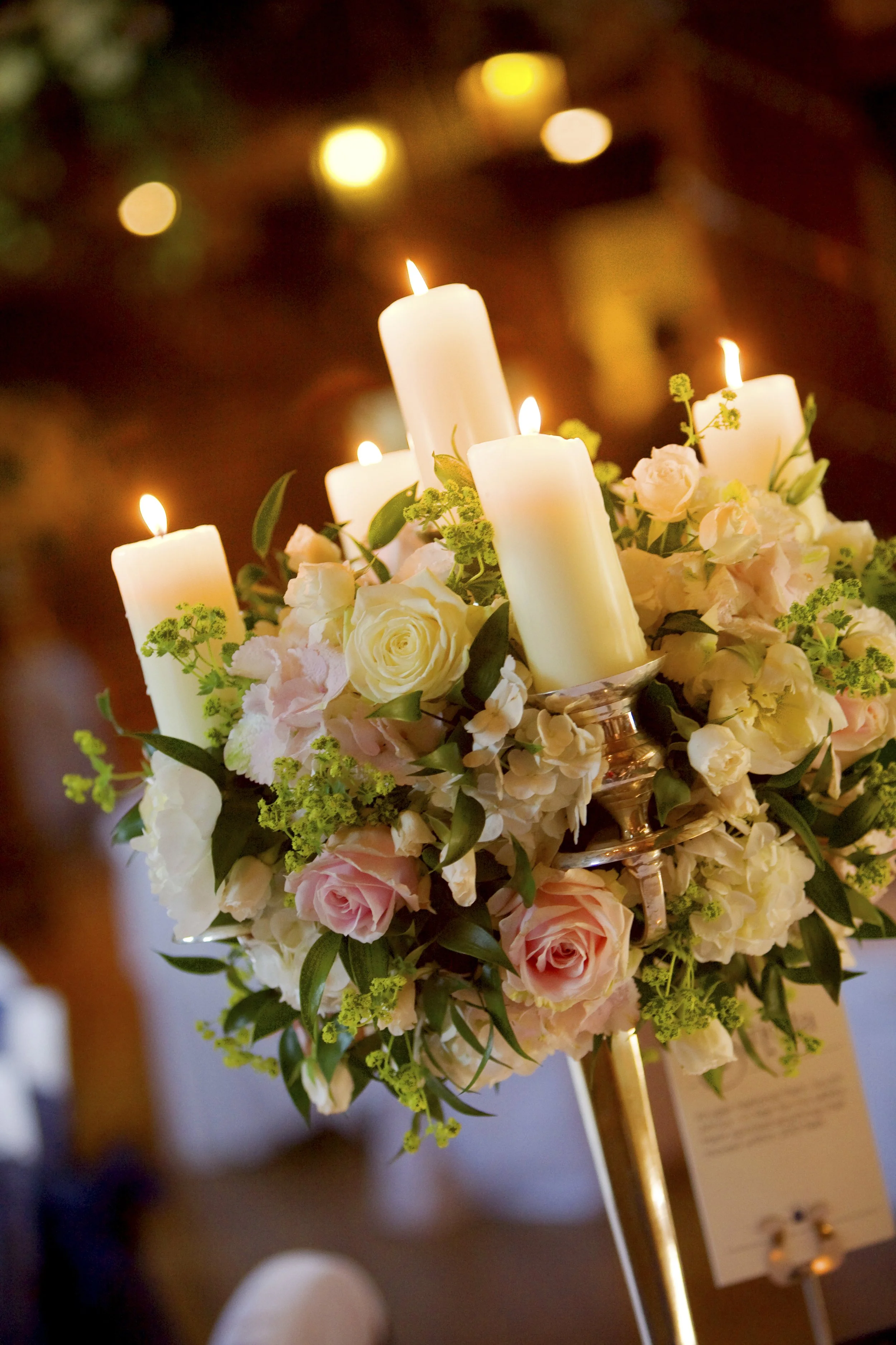 A floral centerpiece with white and light pink roses, surrounded by white candles, lit and placed in a candle holder.