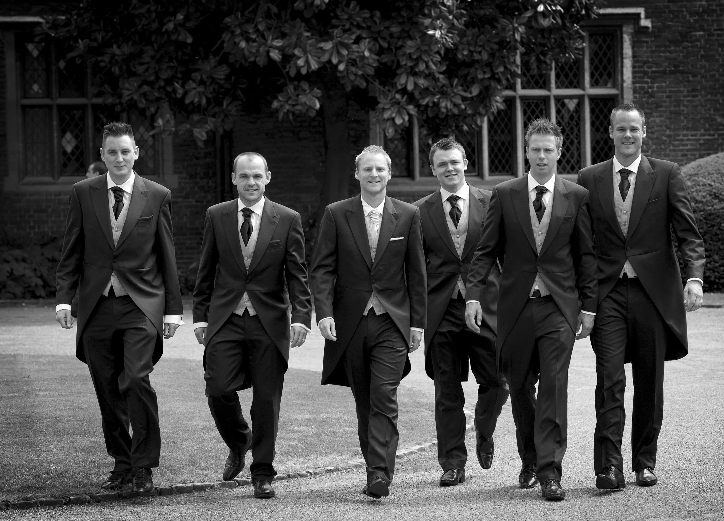 Group of six men in tuxedos walking outdoors in front of a building with large windows and a tree
