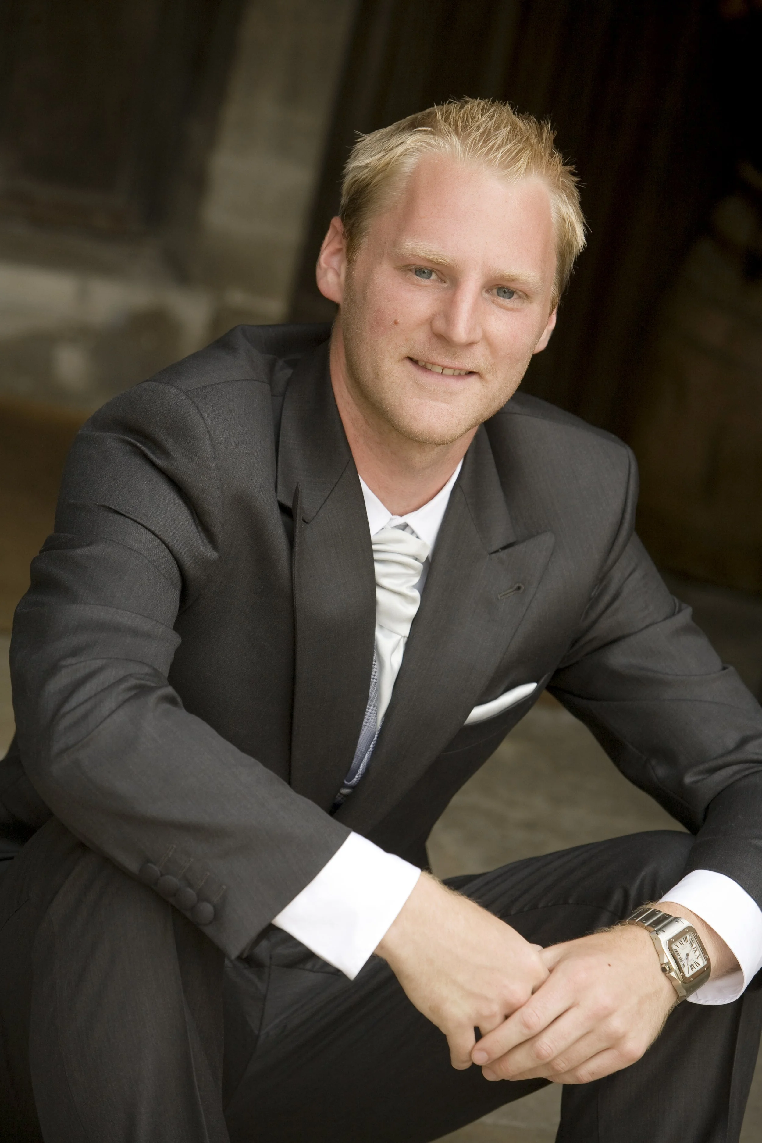 A man in a dark suit with a white dress shirt and a watch, sitting and smiling.