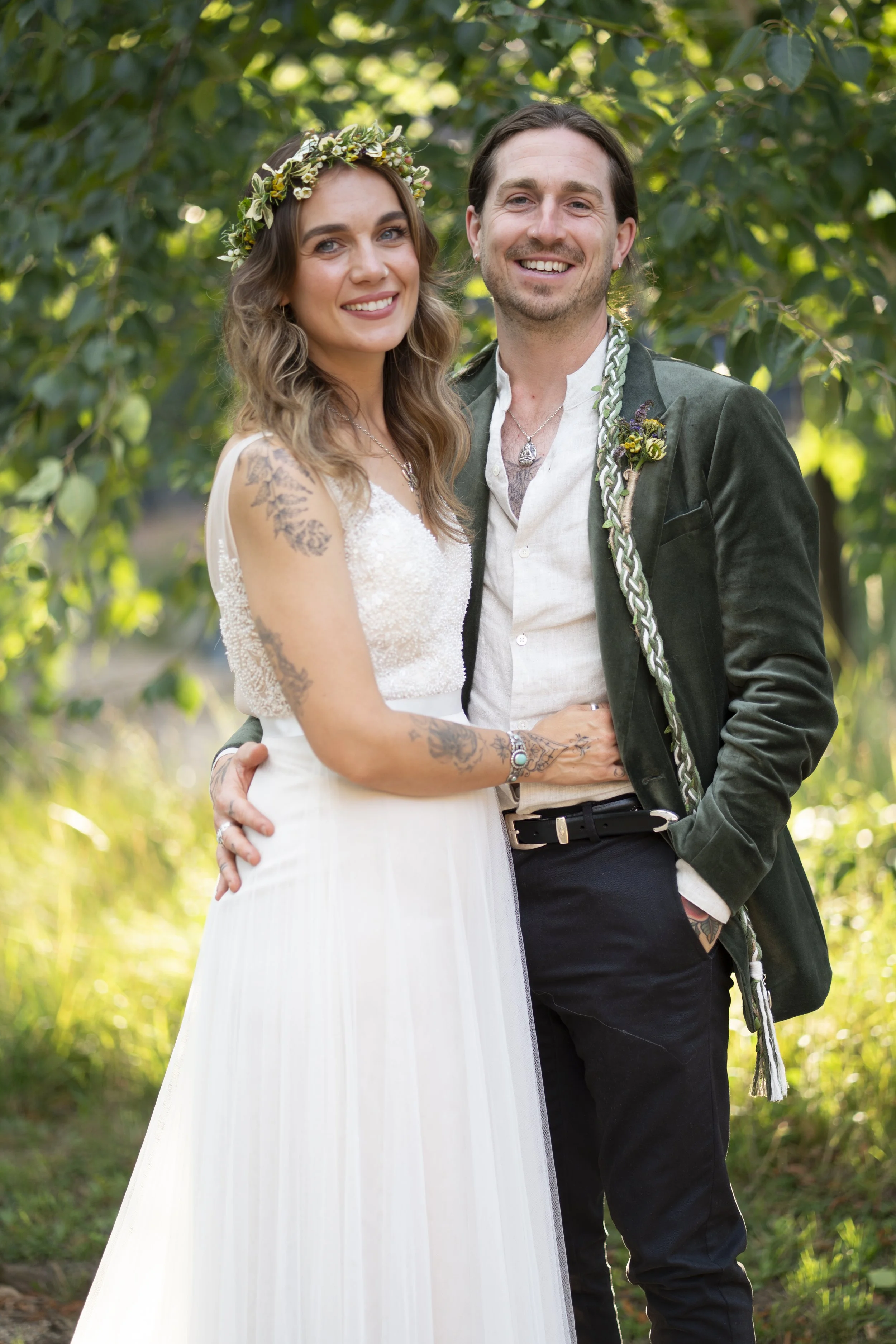 A happy couple poses outdoors on their wedding day, surrounded by green foliage. The bride has long, wavy brown hair, tattoos on her arms, and is wearing a white gown with a floral crown. The groom has dark, shoulder-length hair, and is dressed in a 