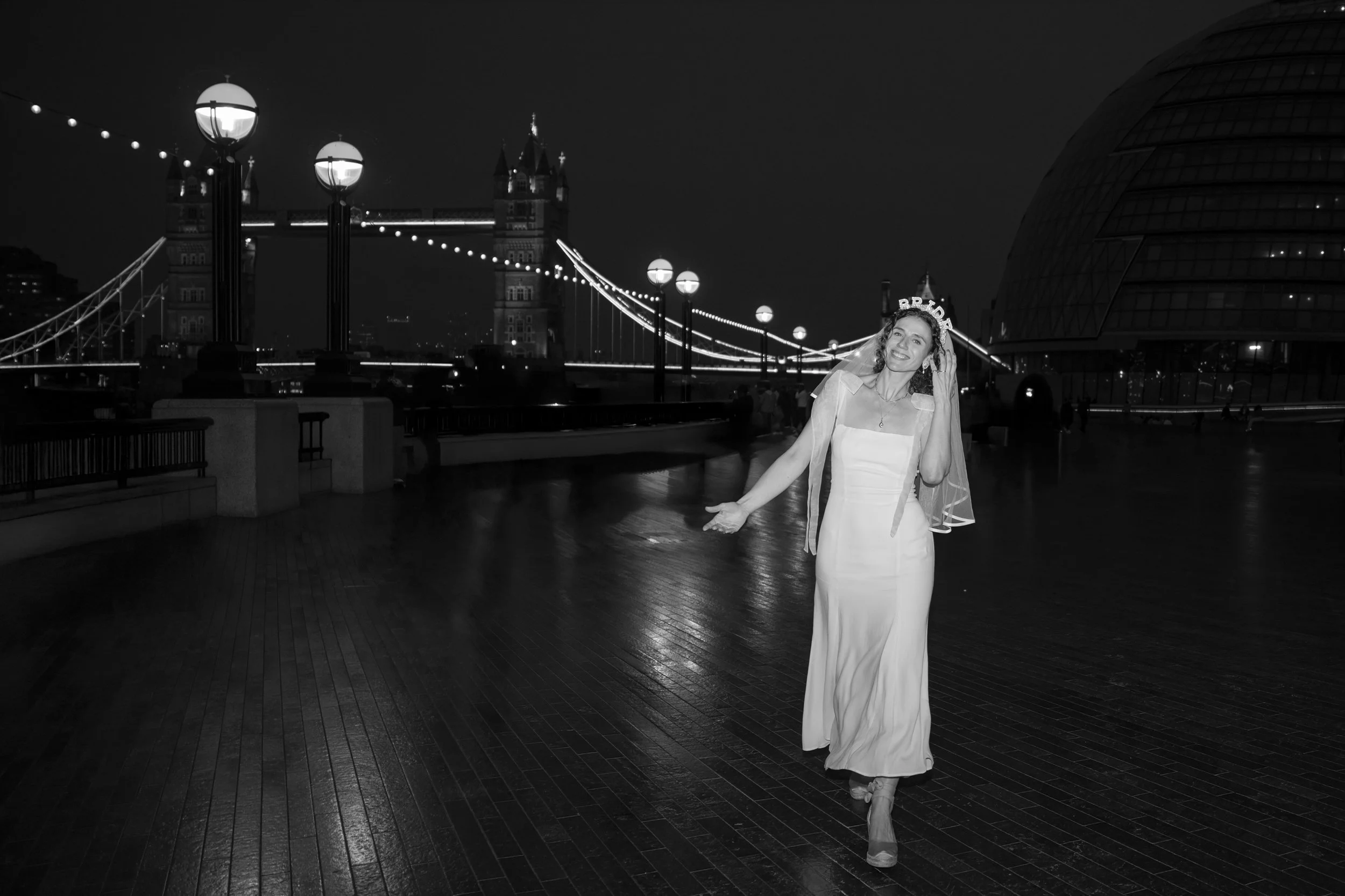 A woman in a long white dress and veil with a tiara walks on a wet wooden walkway at night, with the Tower Bridge and City Hall illuminated in the background.