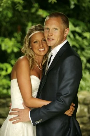 A bride and groom in wedding attire embracing outdoors with green foliage in the background.