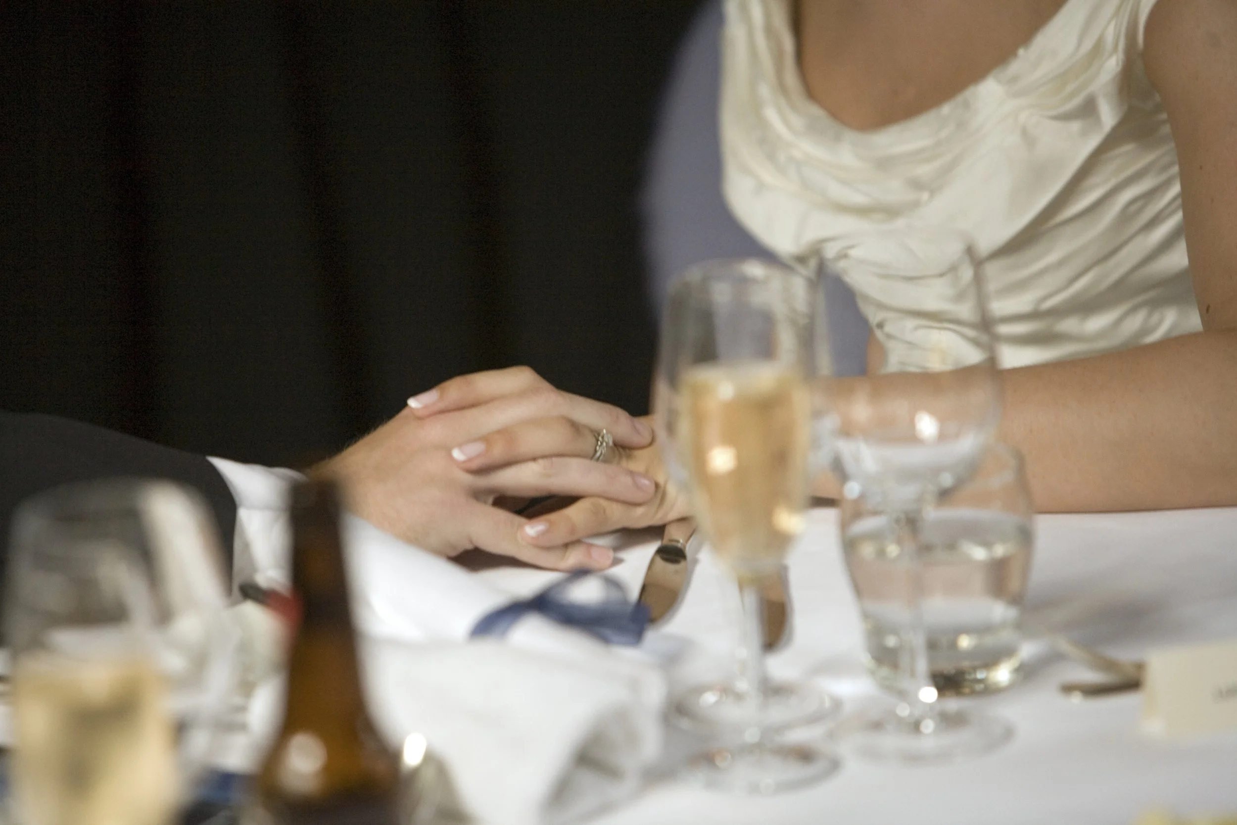 Close-up of a couple holding hands during a wedding reception, with champagne glasses and a bottle on the table.