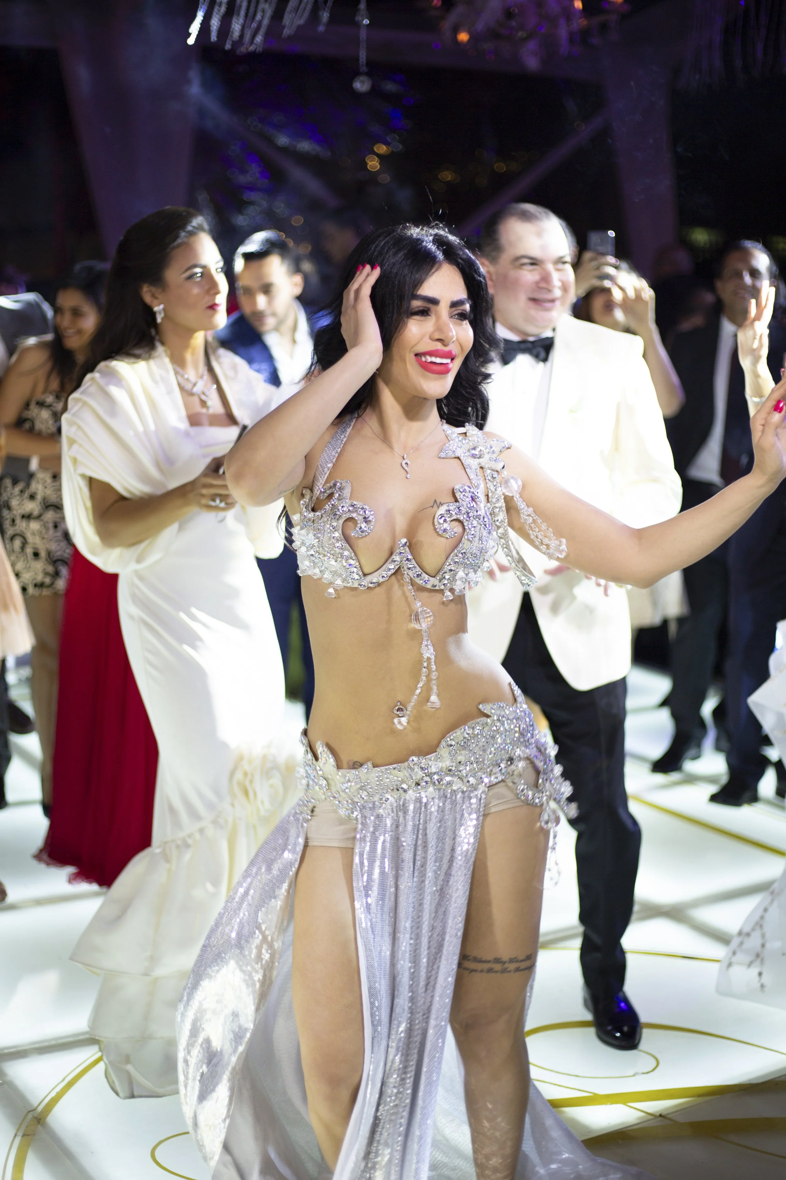 A woman in a silver, jeweled, revealing costume dances at a celebration or party, with other formally dressed people in the background.