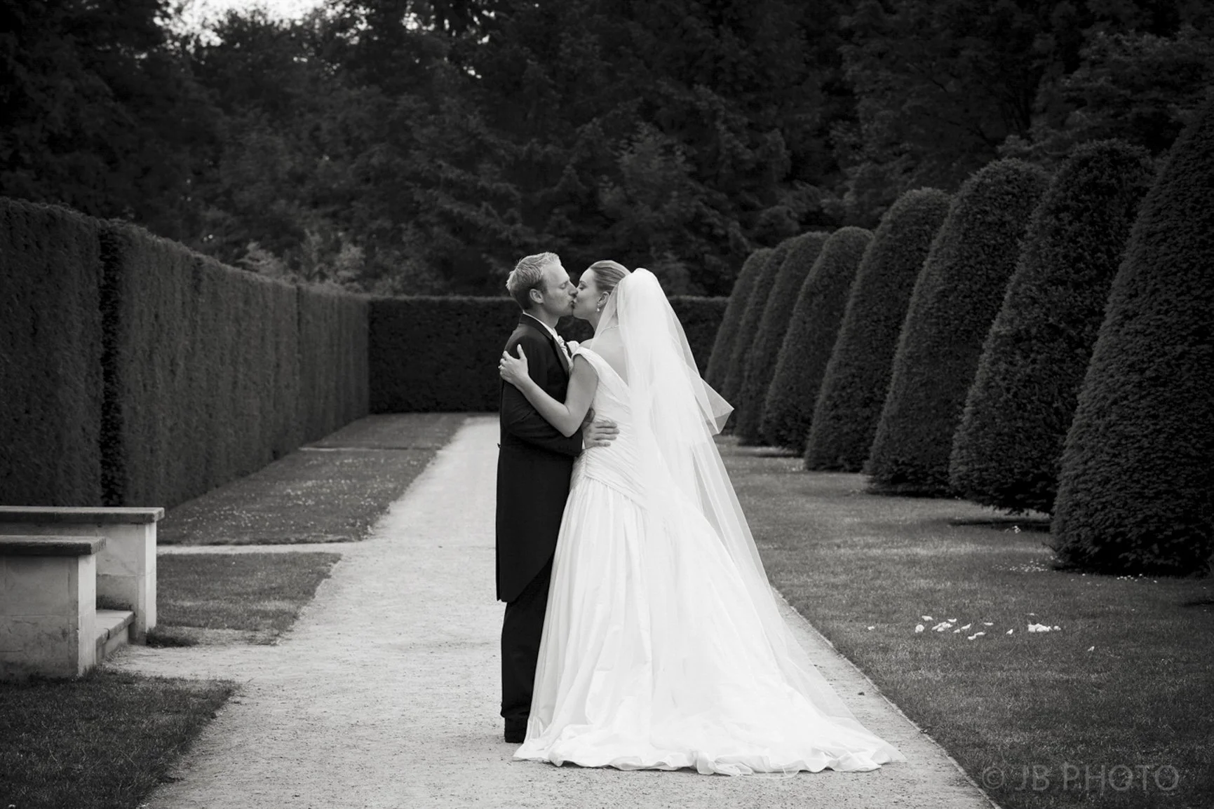 Black and white photo of a bride and groom sharing a kiss on a garden pathway, surrounded by tall, manicured bushes.