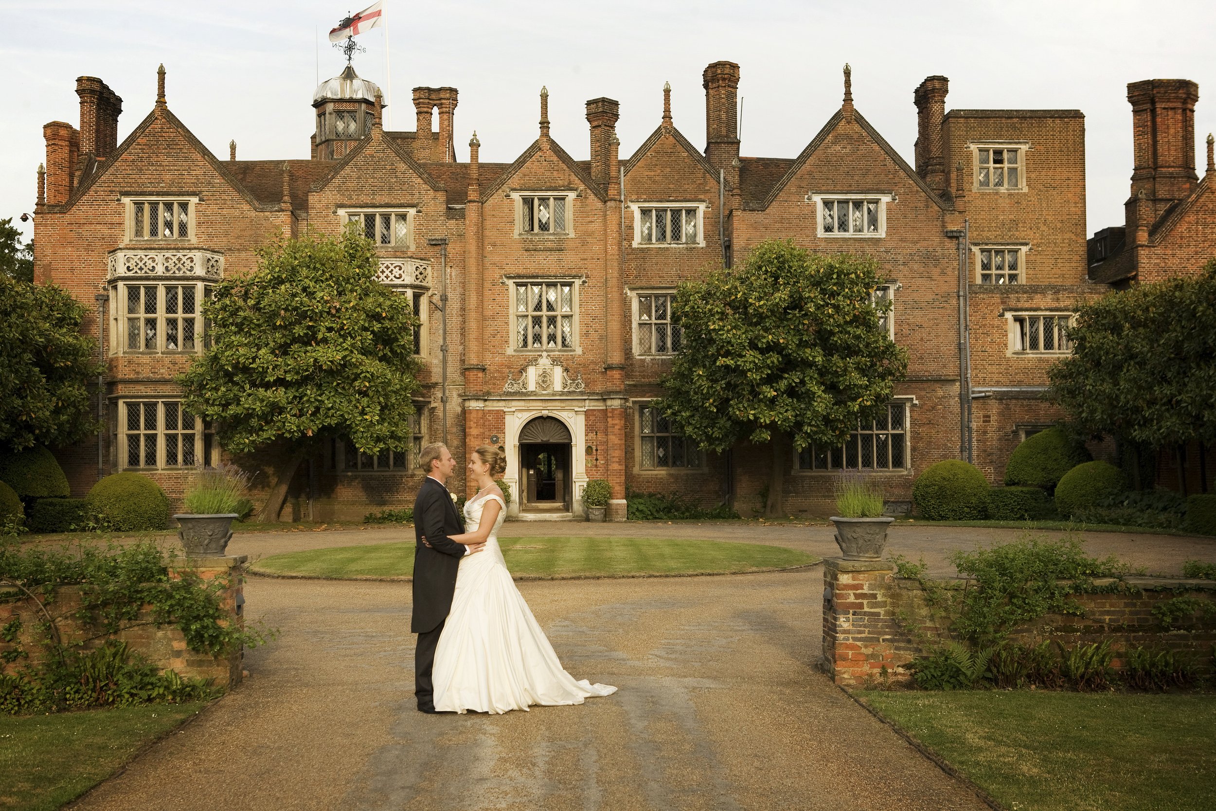 A bride and groom in wedding attire standing in front of a historic brick mansion, holding hands and looking at each other, surrounded by greenery and trees.