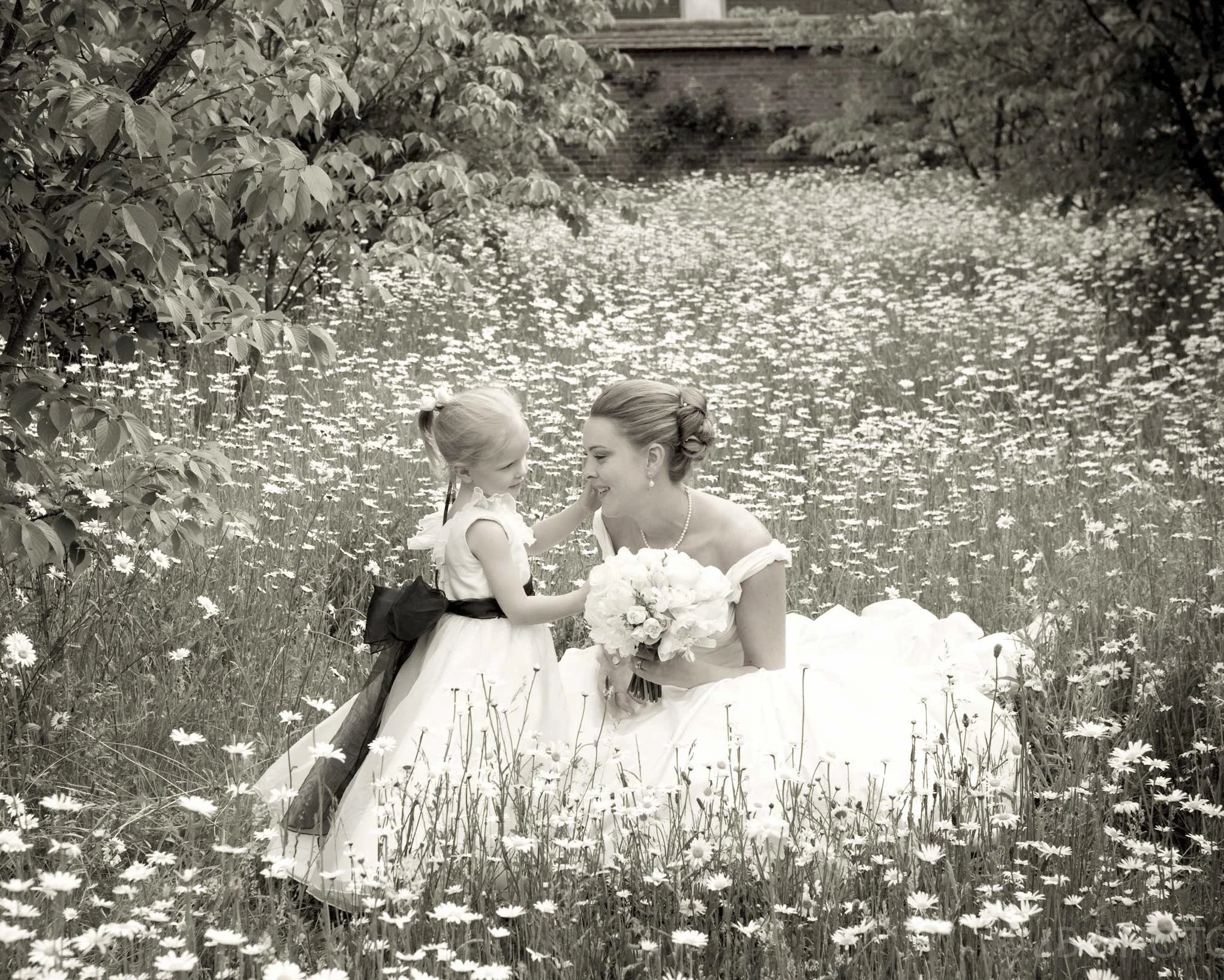 A woman in a wedding dress holding a bouquet of flowers is kneeling in a field of daisies, smiling at a young girl in a flower girl dress with a bow in her hair, as they touch noses amidst a scenic outdoor setting with trees and a small lake in the b