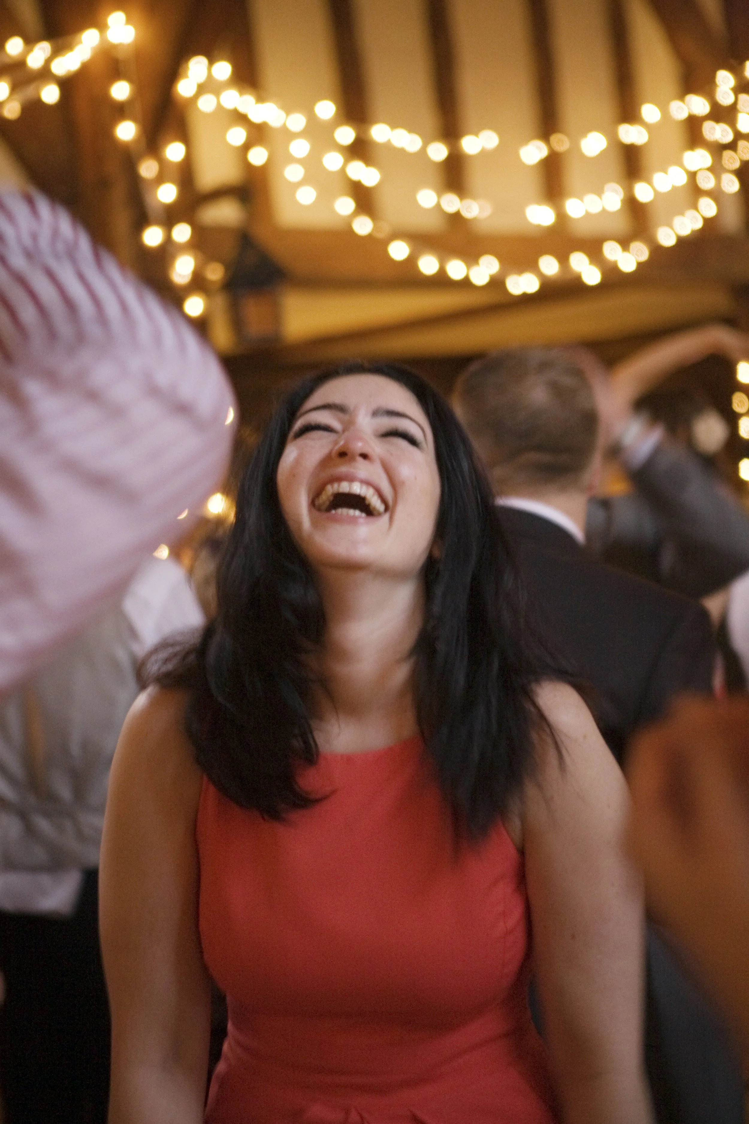 A woman with black hair wearing an orange dress is laughing joyfully at a party or celebration, with string lights and other people in the background.