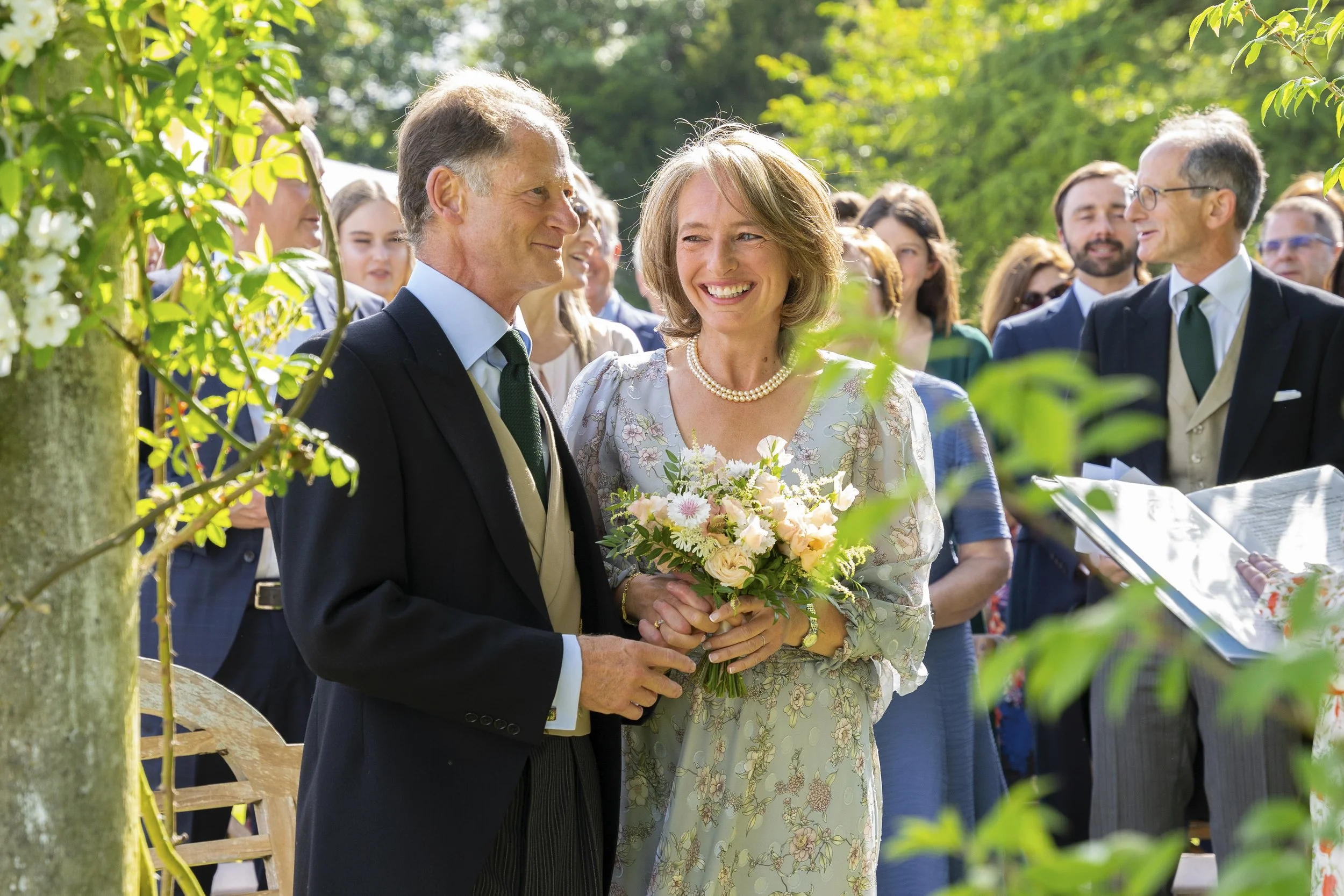 A woman in a floral dress holding a bouquet of flowers is smiling at a man in a suit at an outdoor wedding ceremony. Guests are standing around under green trees, some holding papers, on a sunny day.