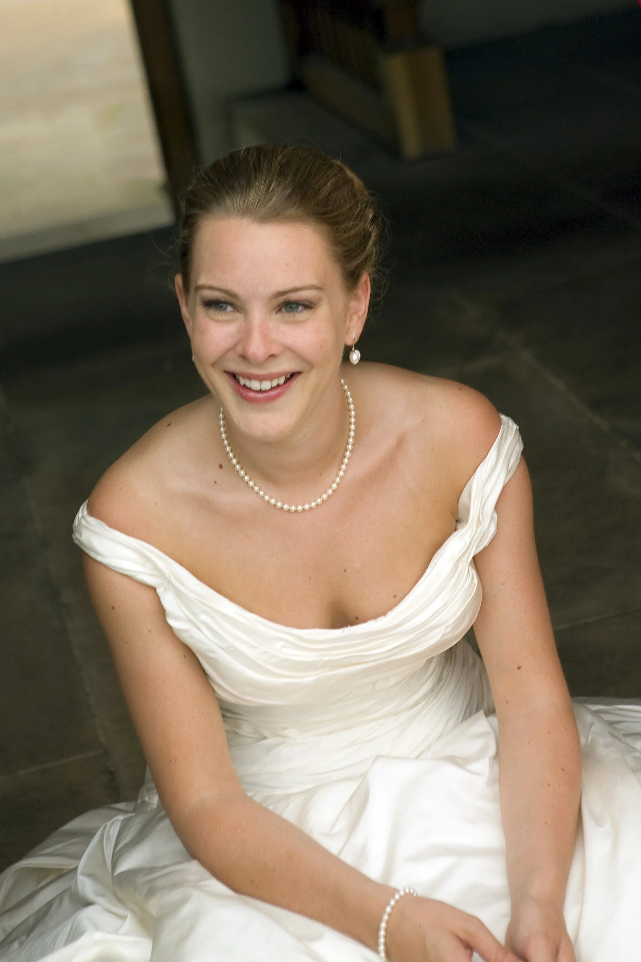 A smiling woman in a white wedding dress, pearl necklace, matching earrings, and bracelet, sitting on the floor indoors.