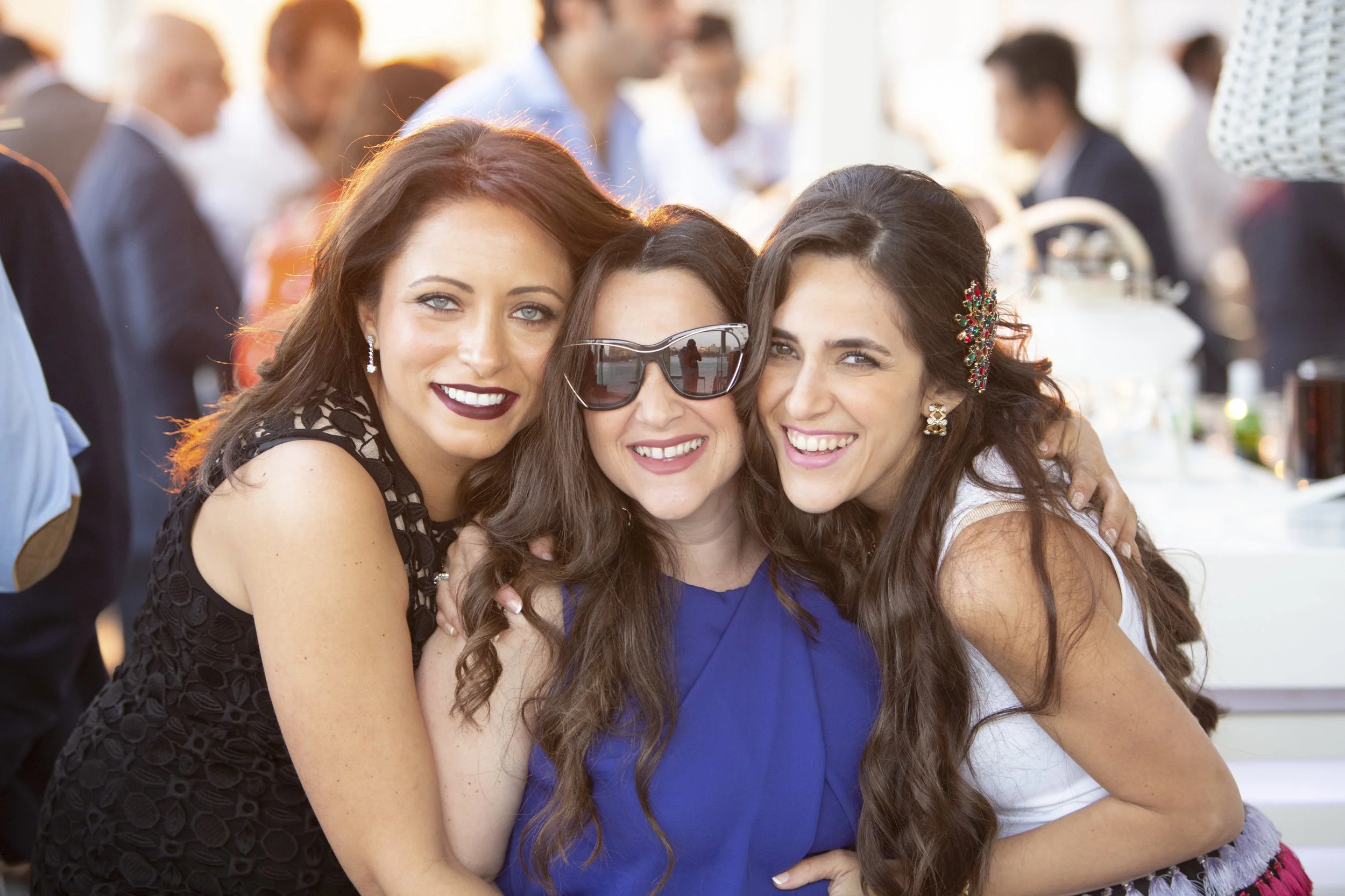 Three young women smiling and hugging at a social gathering or party, with one wearing sunglasses and all in colorful dresses.
