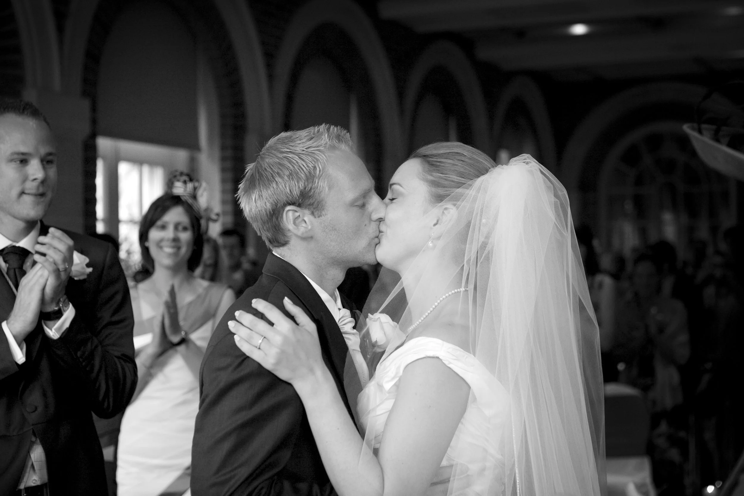 A bride and groom sharing a kiss during their wedding ceremony, with guests clapping in the background.