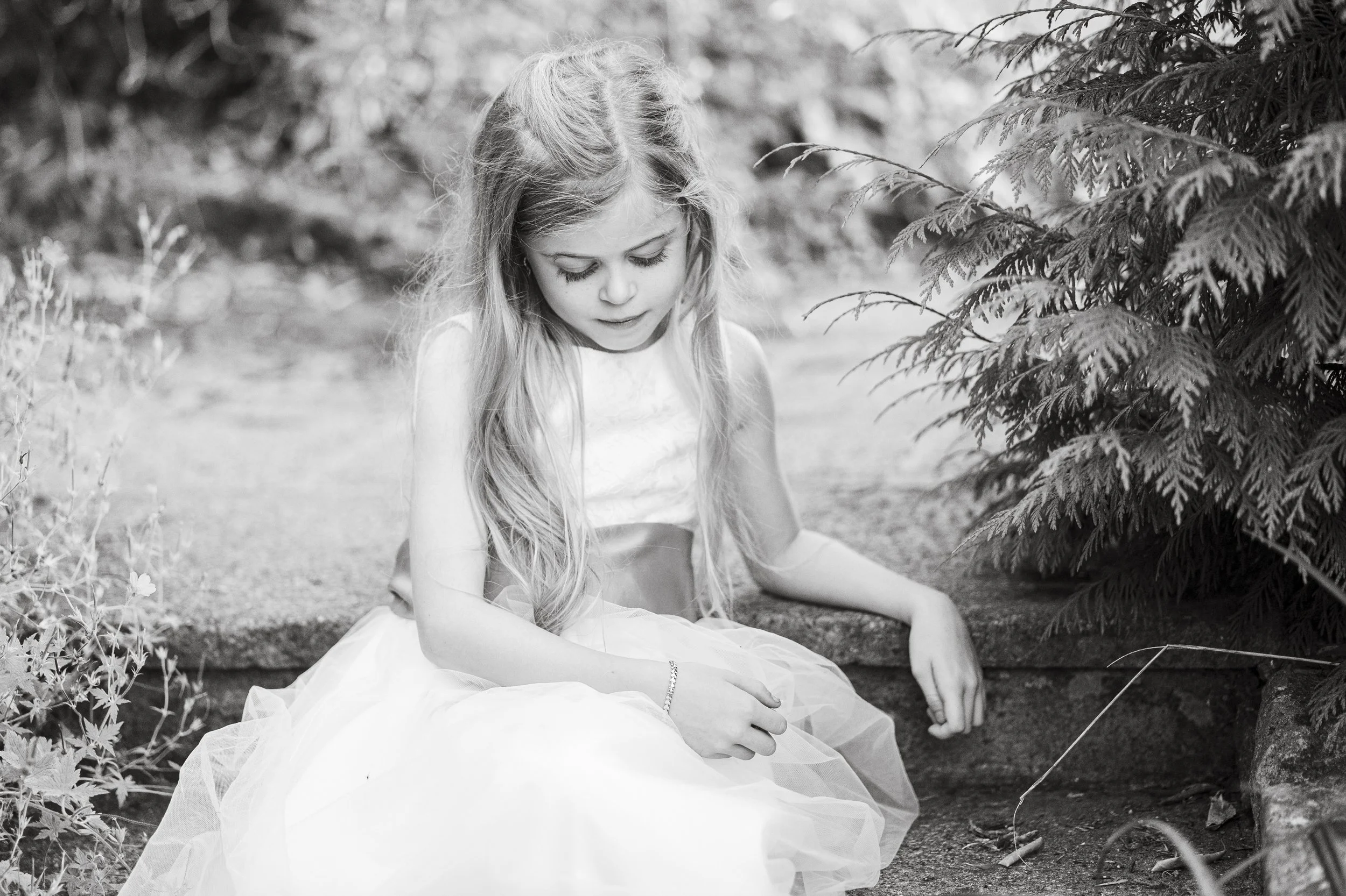 A young girl in a white dress sitting on the ground near plants and bushes, looking down.