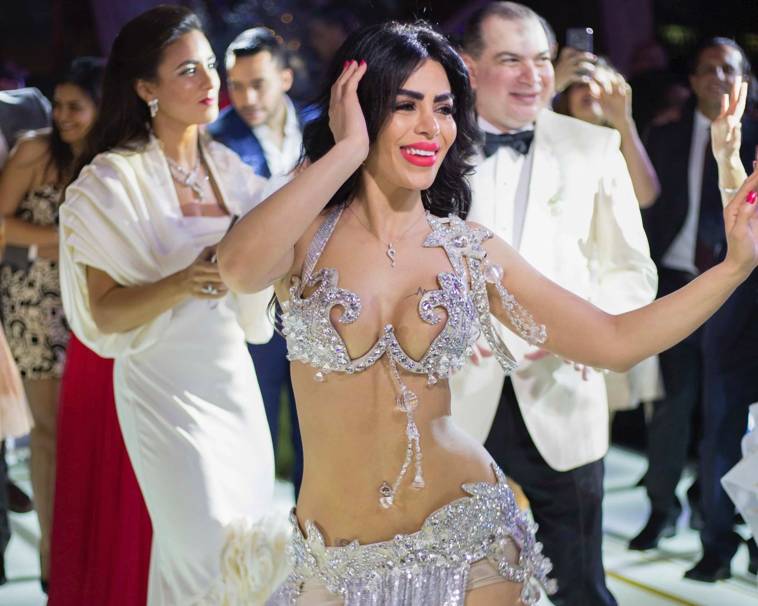A woman in a sparkling, elaborate costume with rhinestones and chains at a formal event, smiling and touching her hair, surrounded by elegantly dressed people.