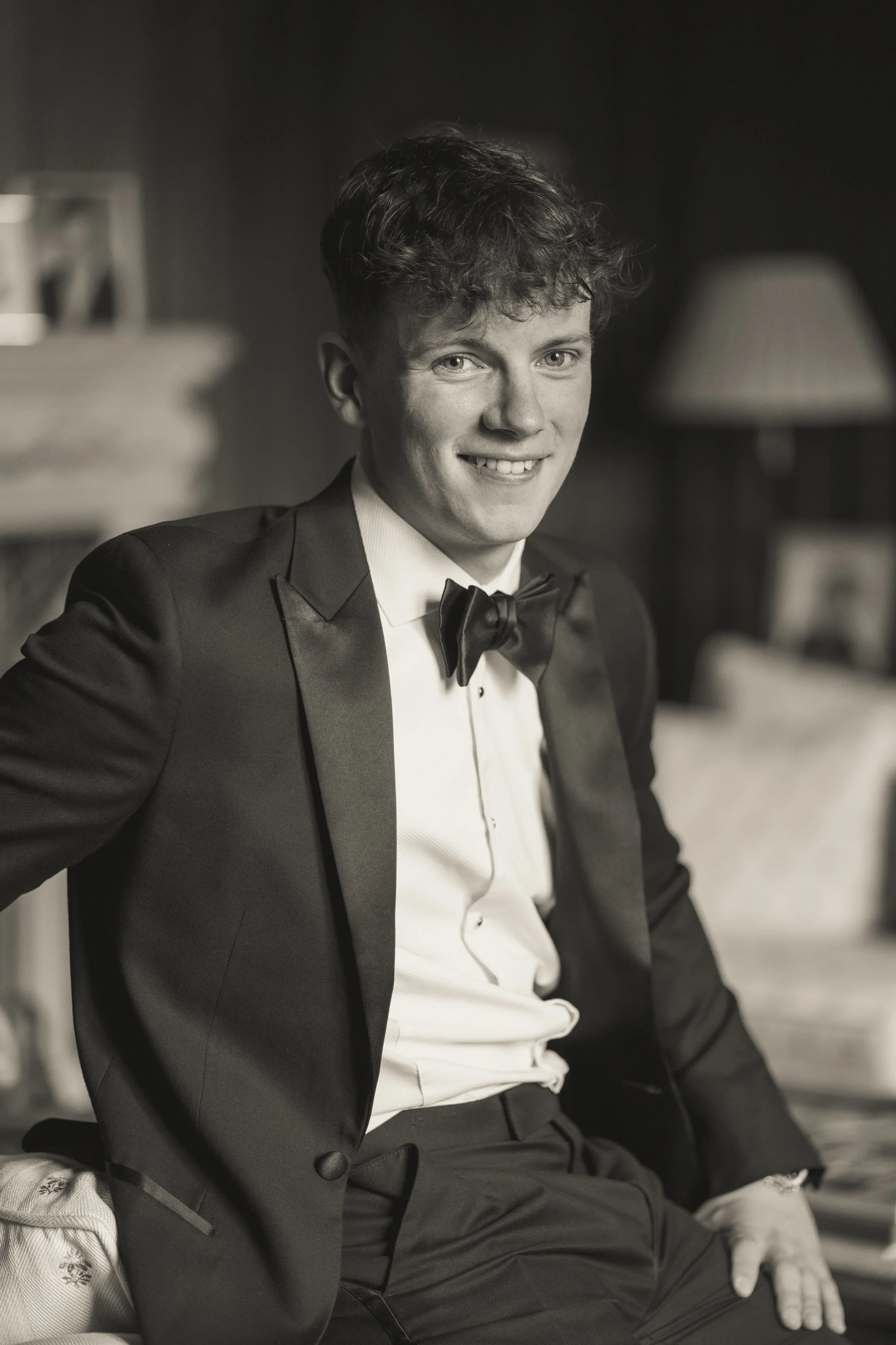 A young man wearing a tuxedo and bow tie, sitting on a bed in a living room, smiling at the camera.