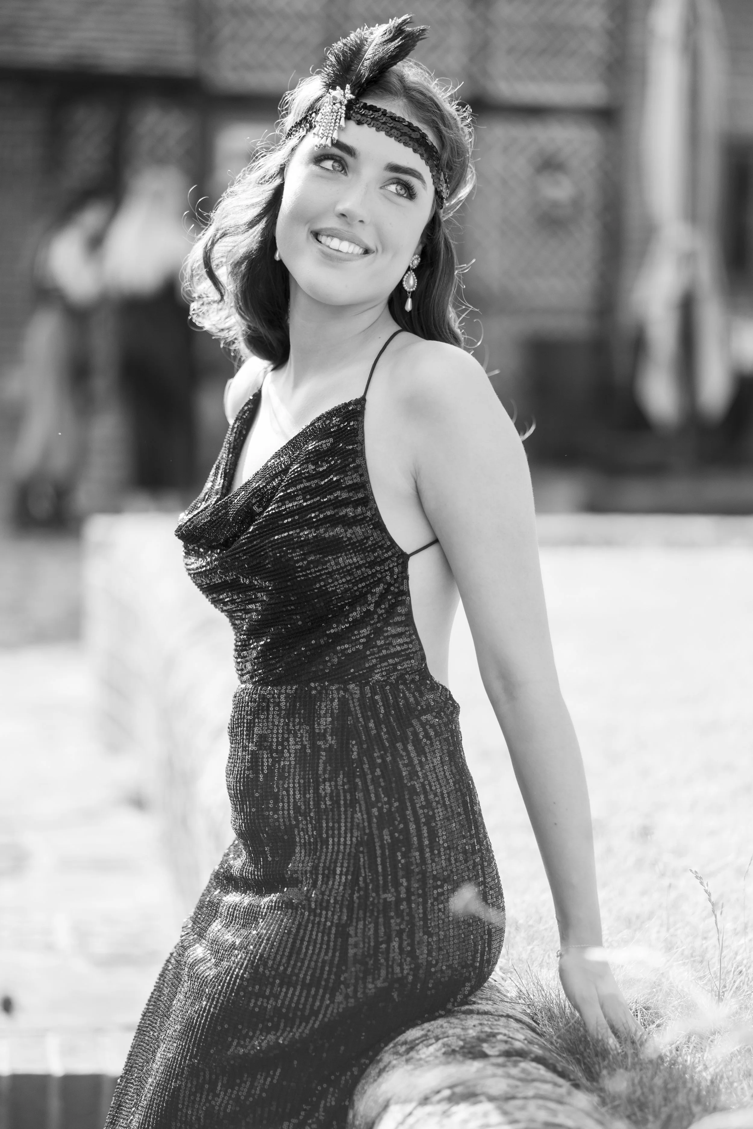 A woman in a sequined dress sitting on a log outdoors, smiling and looking up. She is wearing a feathered headband and earrings.