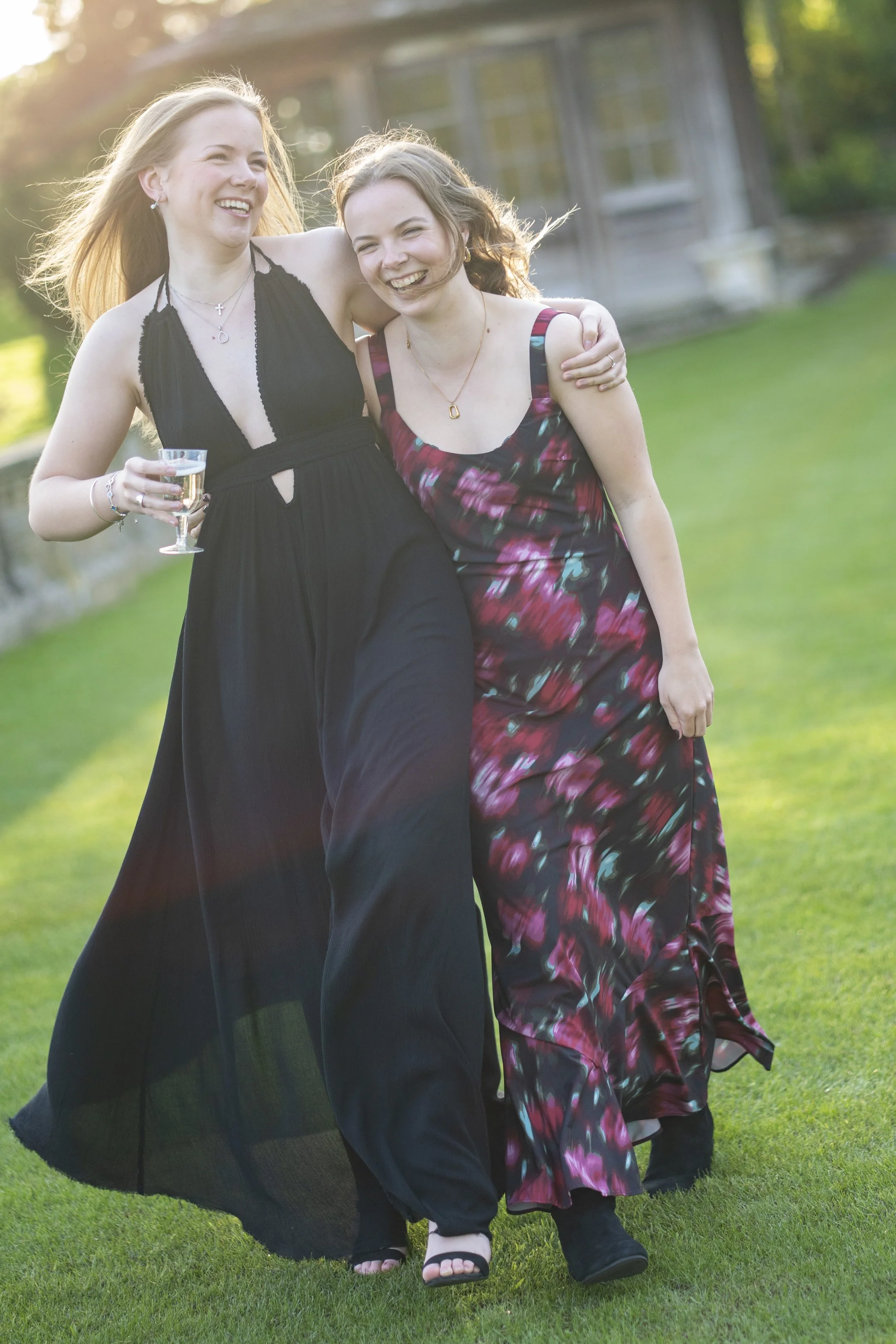 Two young women in elegant dresses smiling and laughing together outside, with arm around each other, one holding a glass of champagne, sunlight streaming in background.