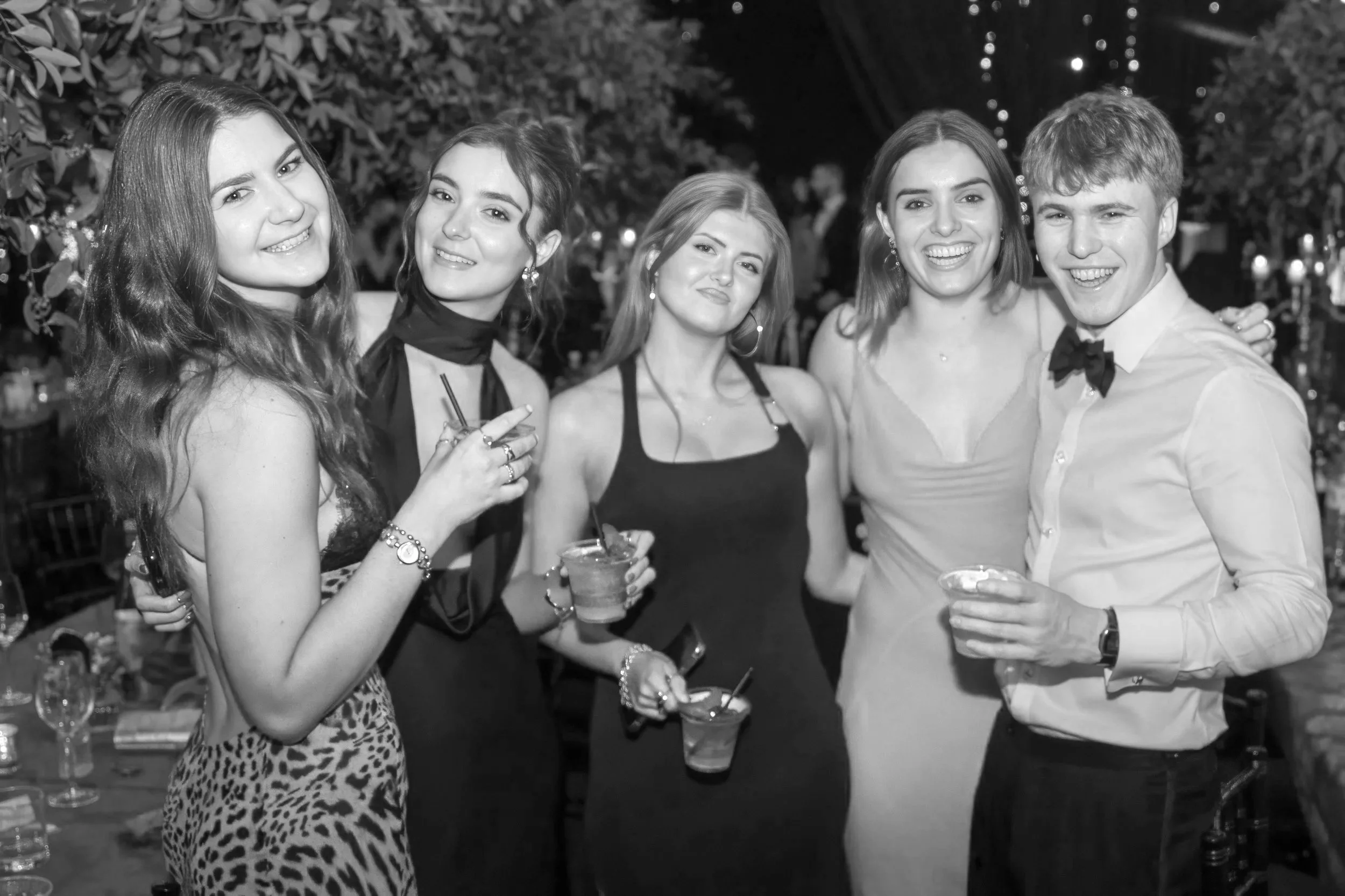 Five young adults, four women and one man, smiling and holding drinks at a party or social gathering in a dimly lit setting.