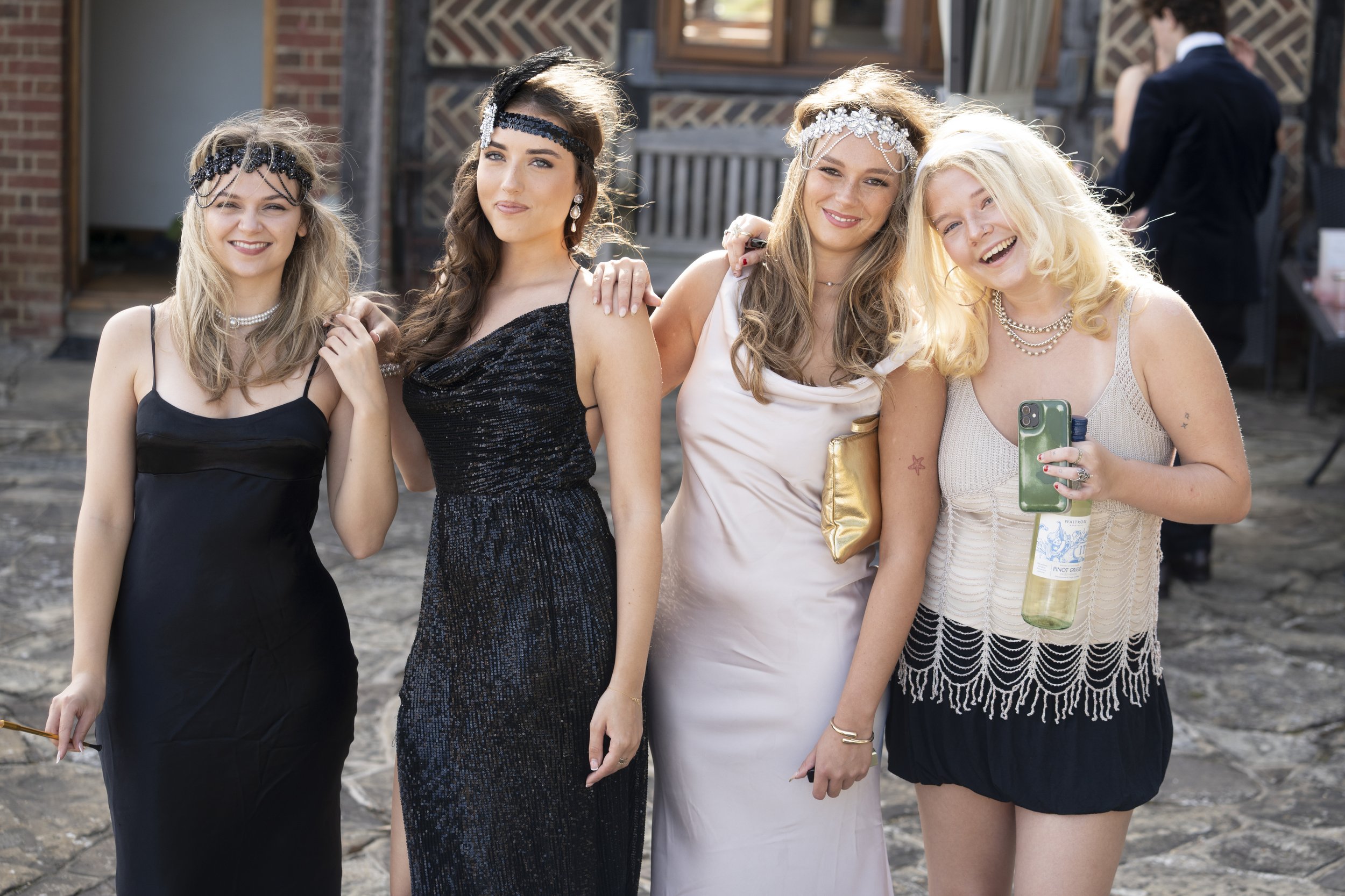 Four women dressed in evening gowns, standing side by side outdoors, smiling at the camera, with a brick building in the background.