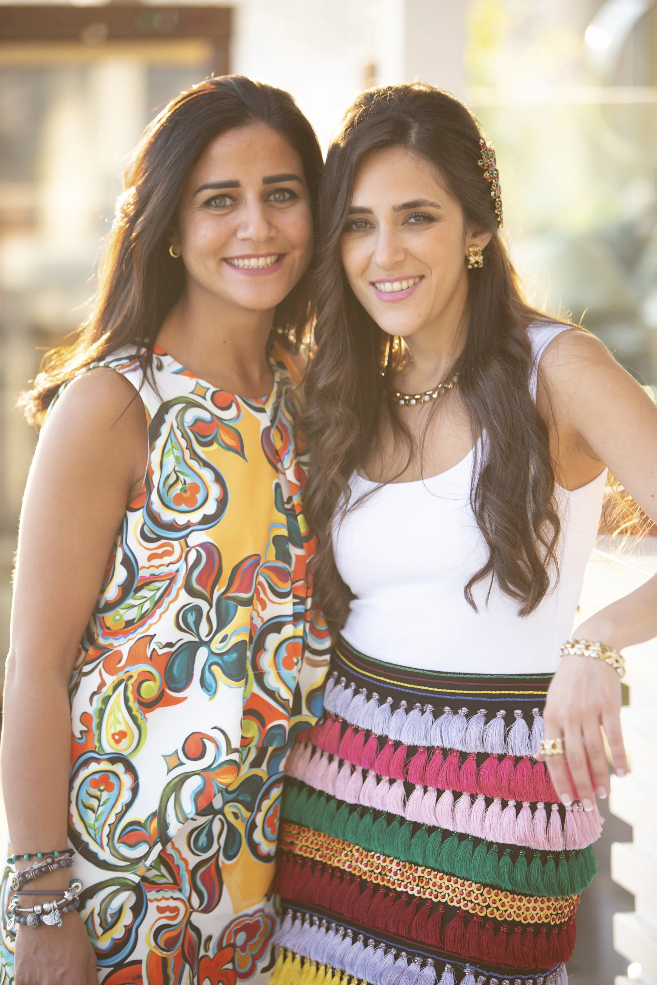 Two women standing close together outdoors, smiling at the camera during a sunny day, with one wearing a colorful patterned dress and the other in a white top and aMulticolored fringed skirt.