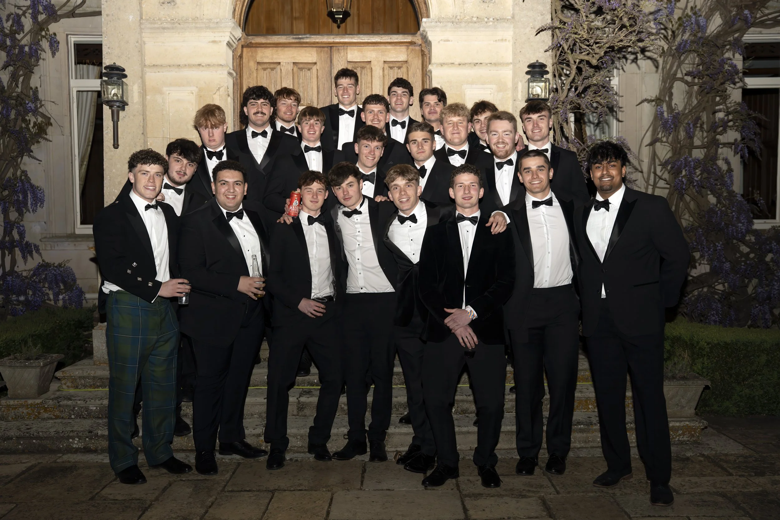 Group of young men dressed in tuxedos and suits, posing for a photo outside a building at night.