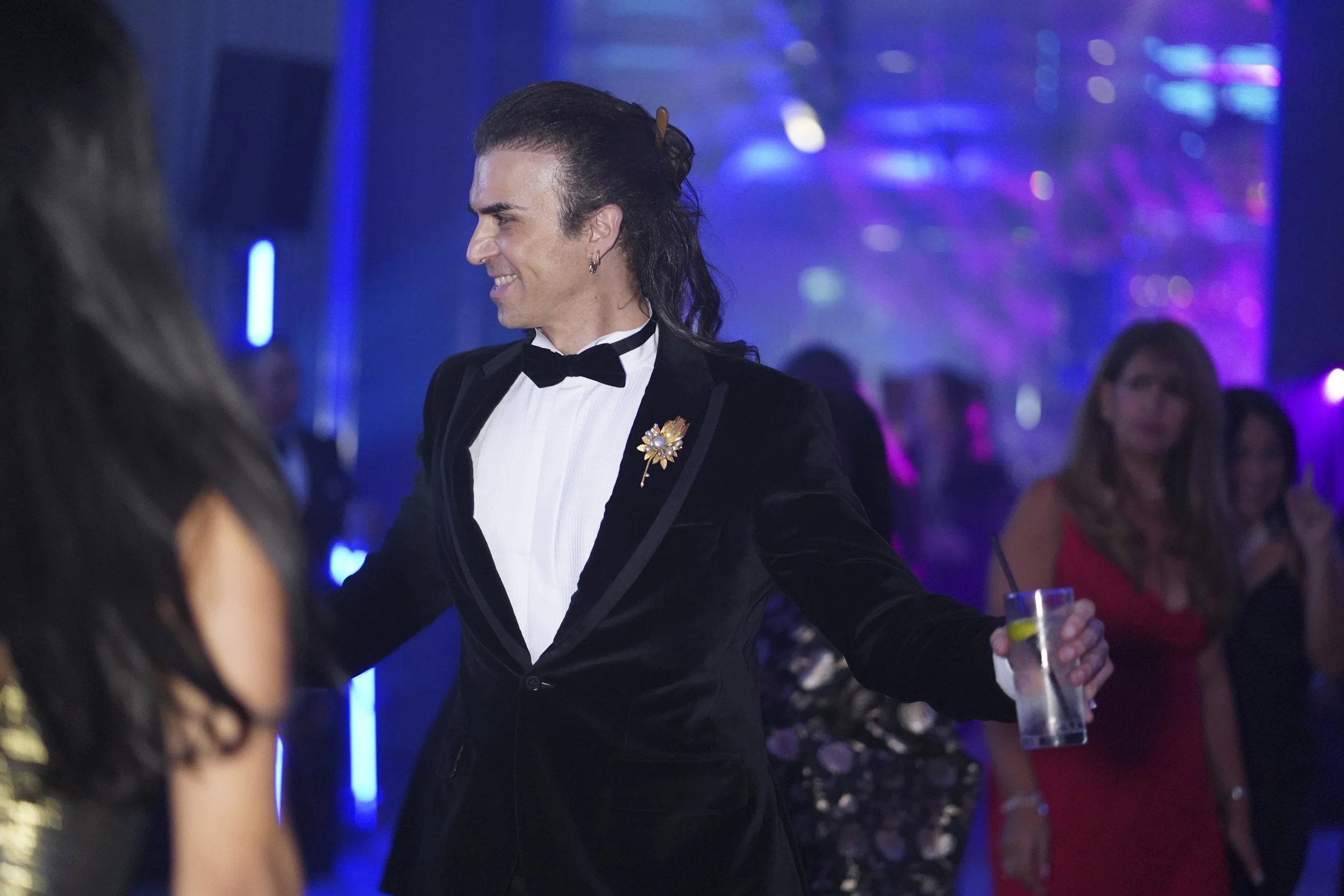 A man in a black tuxedo with a white shirt and black bow tie, holding a drink, smiling and engaging with others at a social event with colorful lighting and other people in the background.