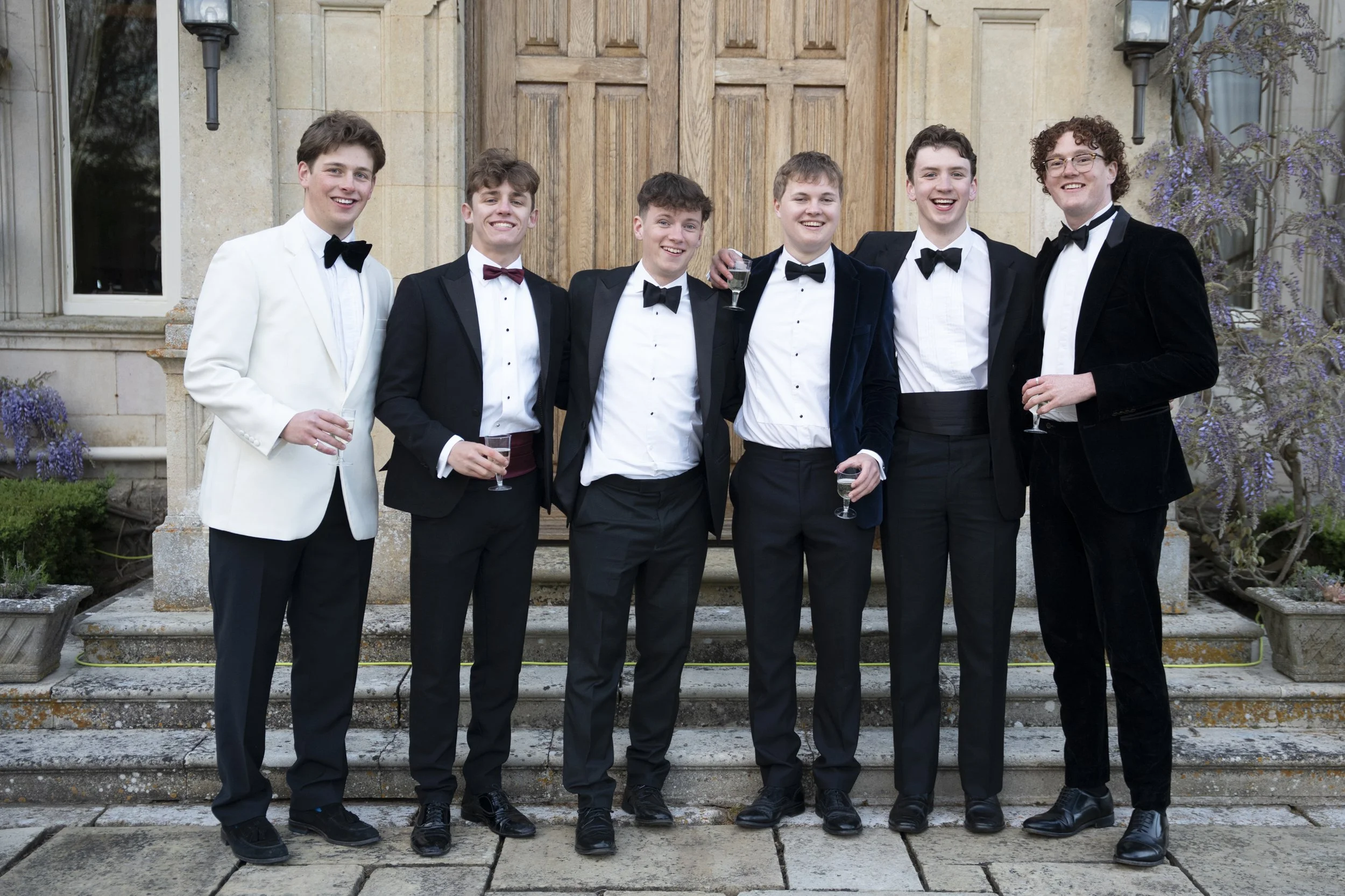 Seven young men in formal tuxedos with bow ties, standing on steps in front of a wooden door and stone building, celebrating with drinks.