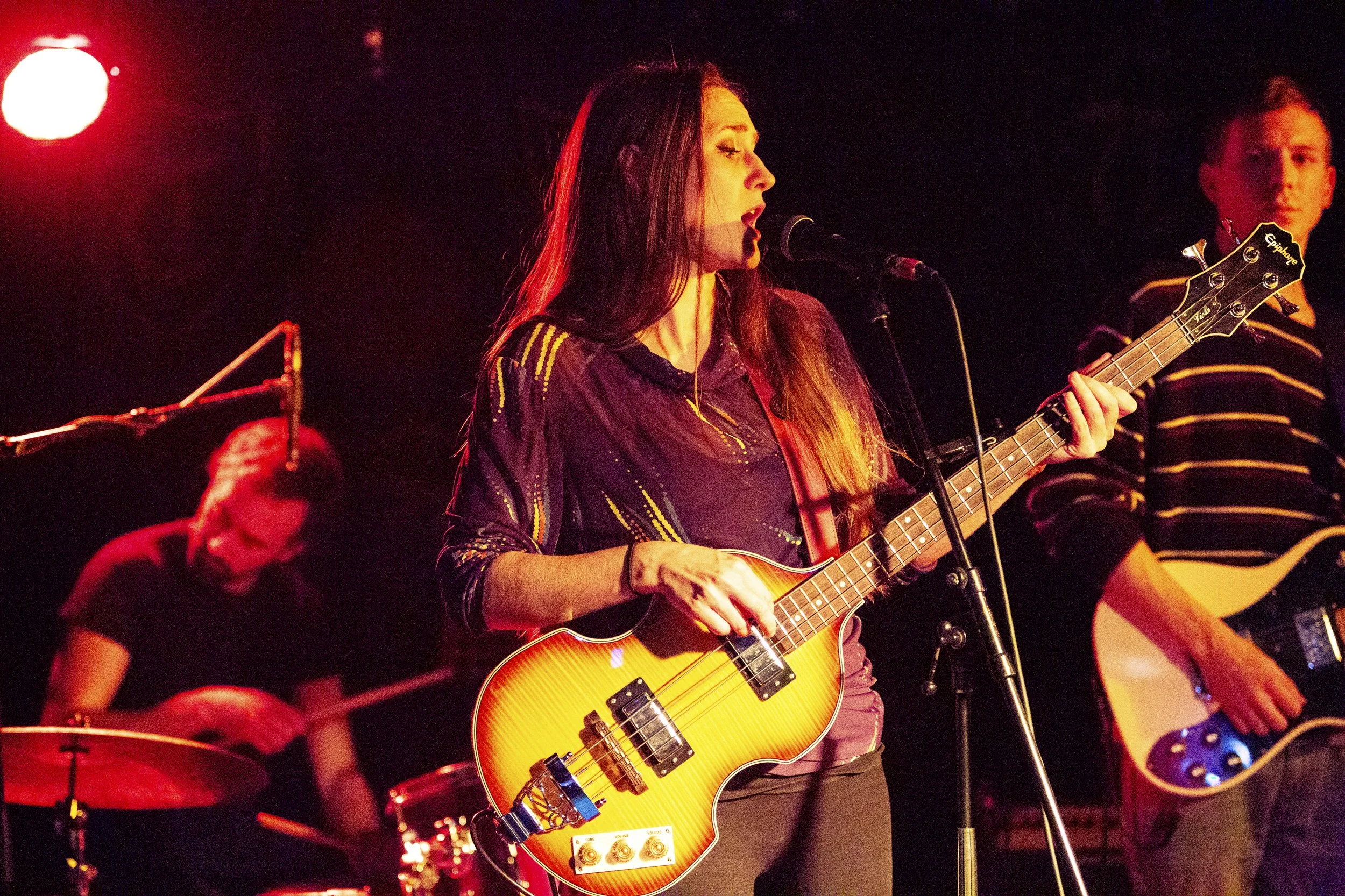 Women playing electric guitar and singing into a microphone on stage with band members, including a drummer in the background, under red stage lighting.