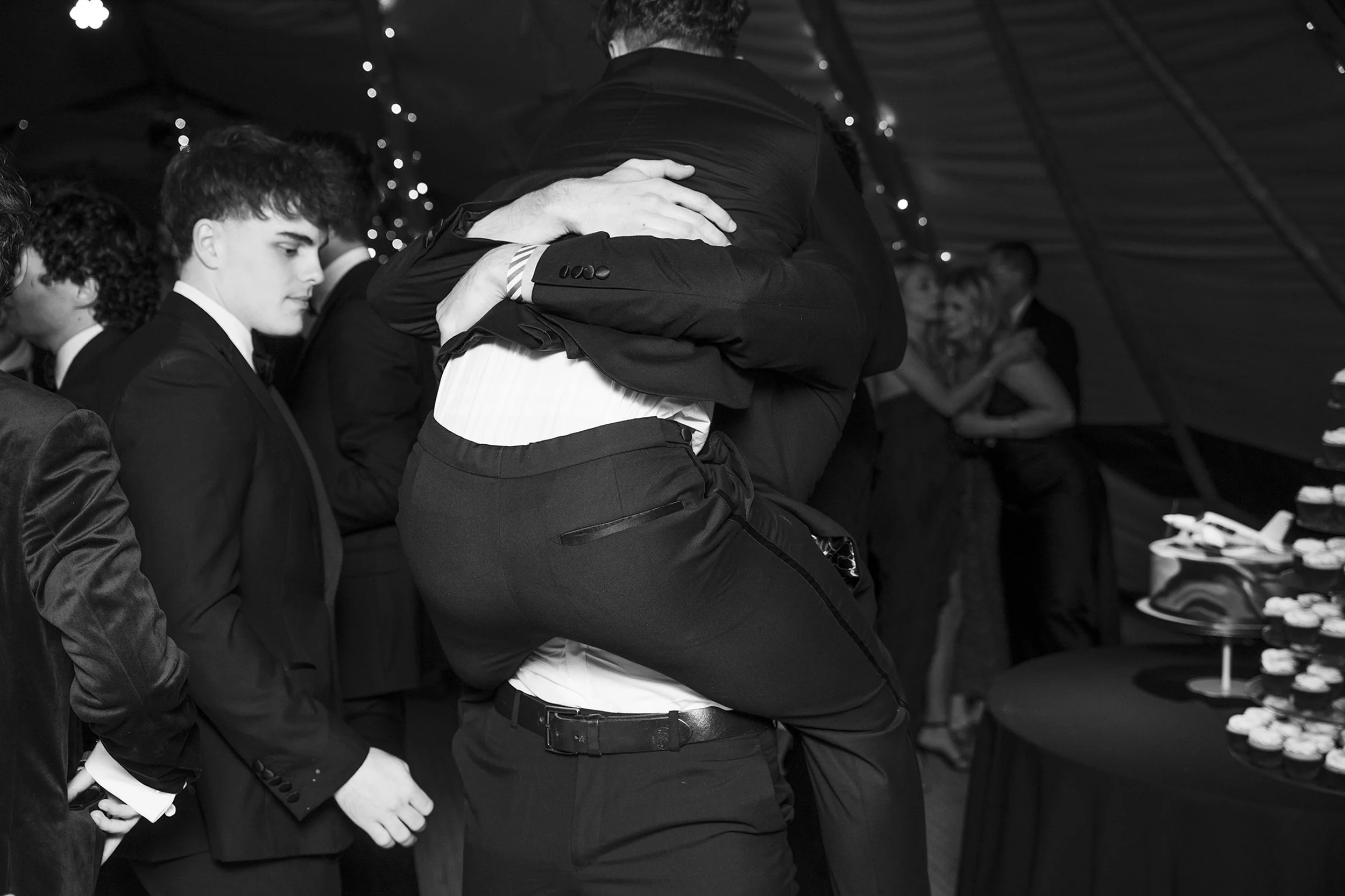People in formal attire at a party, with two individuals embracing in the foreground, and others standing nearby, some smiling in the background, with desserts on a table to the right.