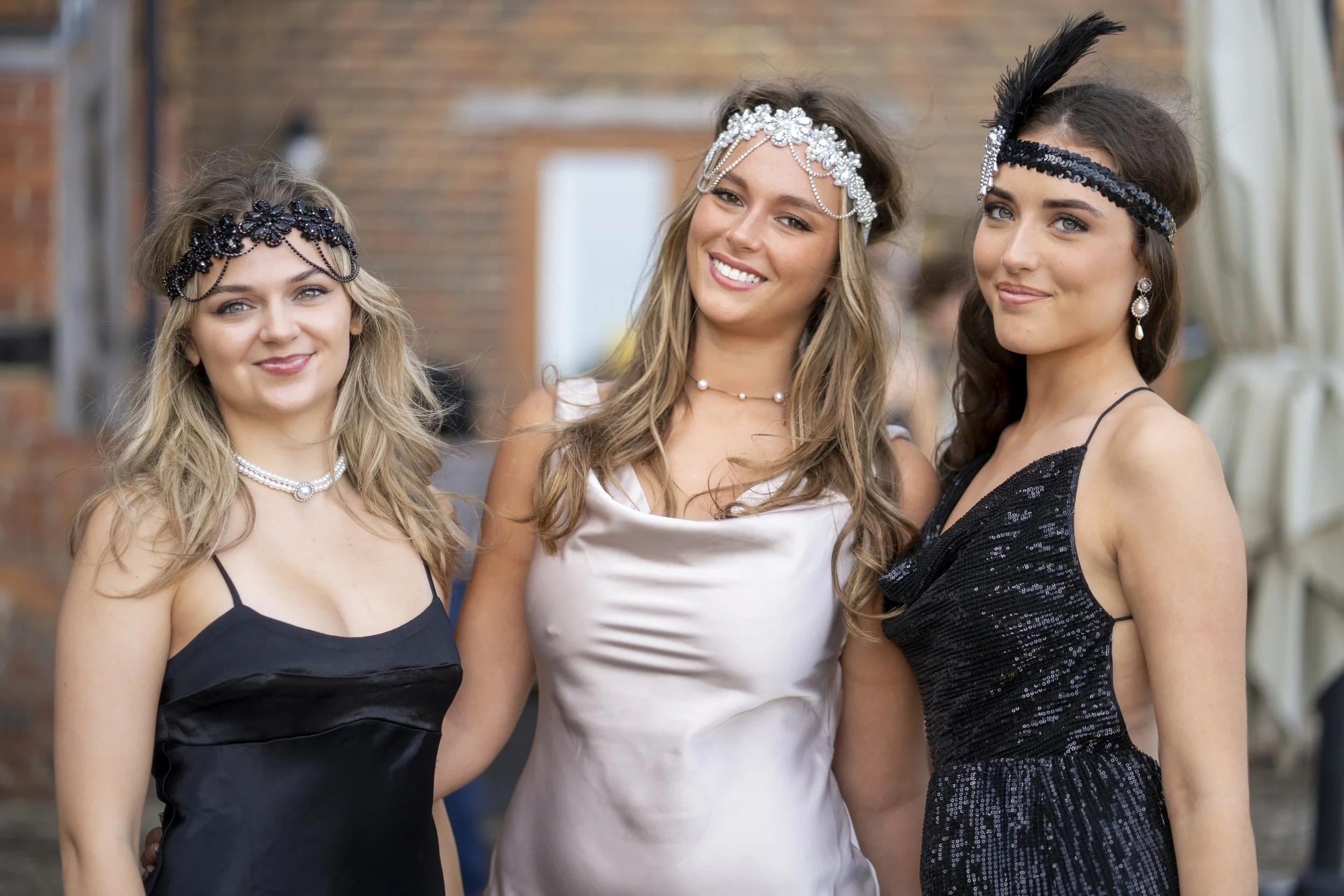 Three young women dressed in elegant evening wear and wearing decorative headbands, standing outdoors and smiling.