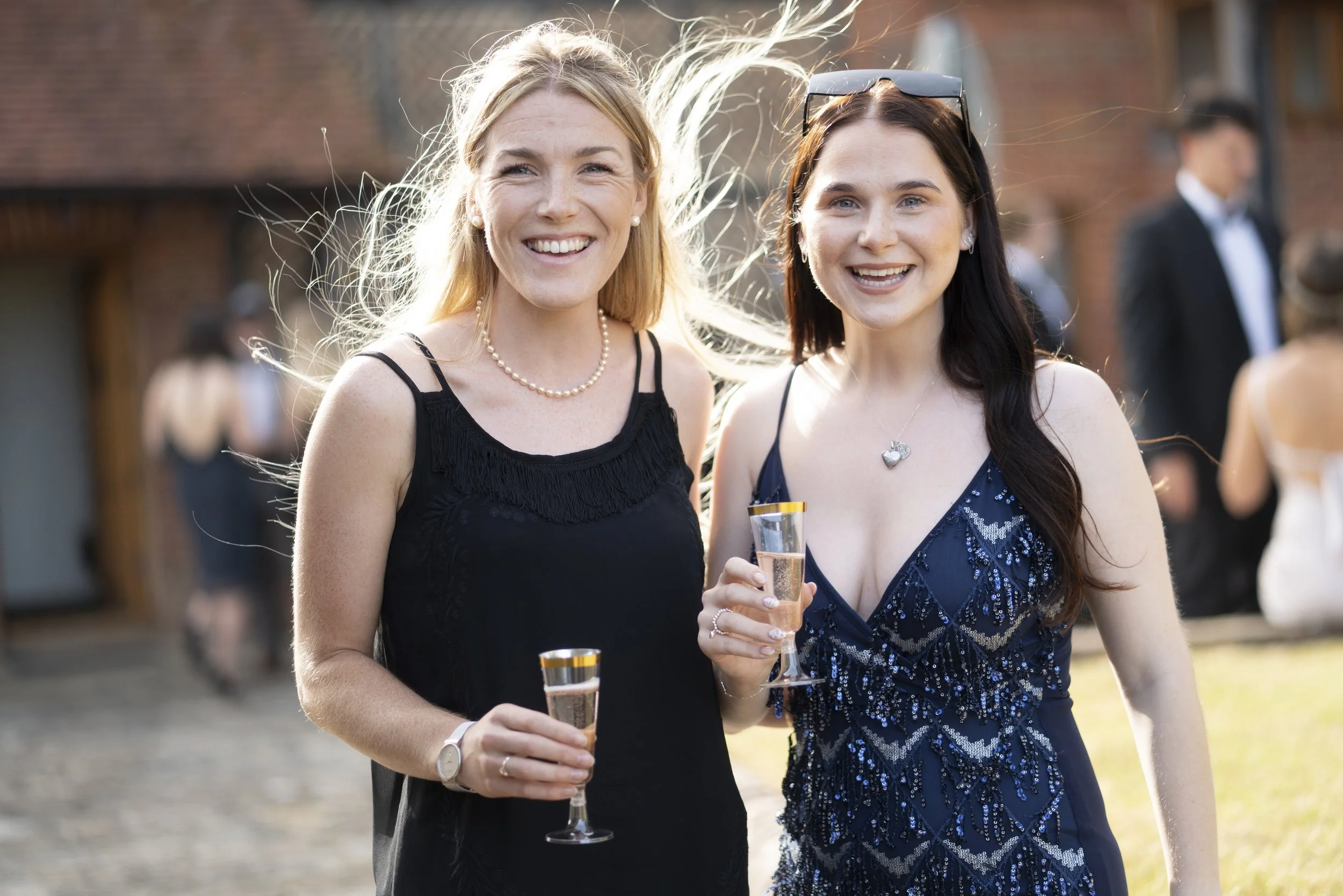 Two women smiling and holding glasses of champagne outdoors at a social event, dressed in elegant summer dresses.