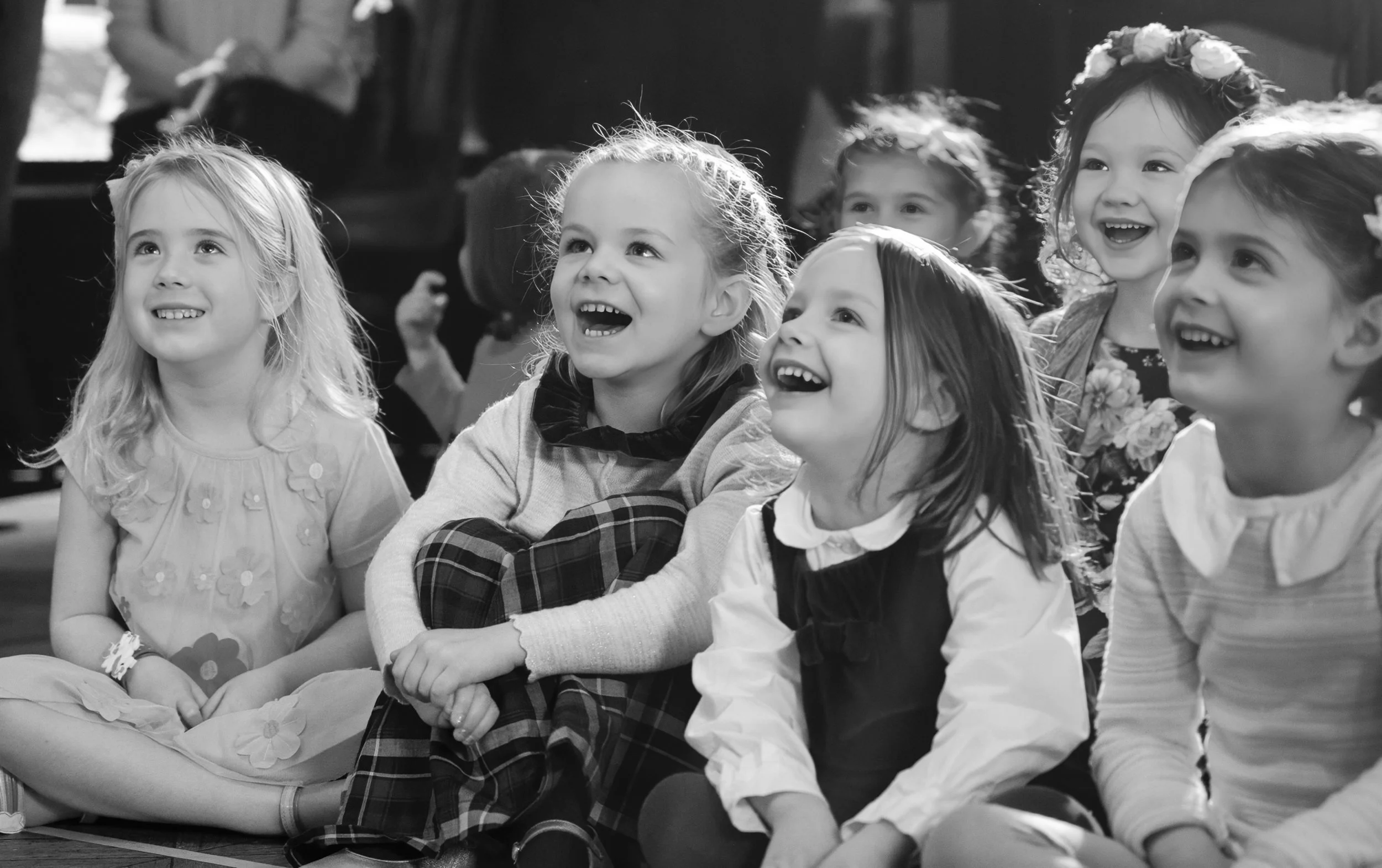 A group of young girls sitting on the floor, smiling and laughing, in a cheerful indoor setting.