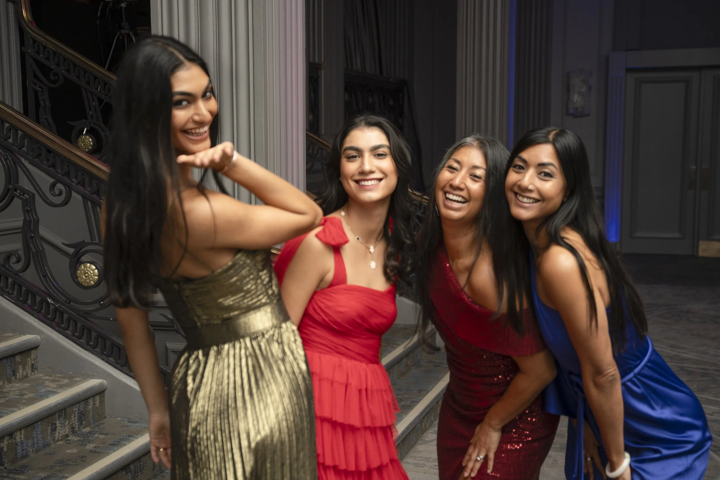 Four women in colorful dresses smiling and posing together on a staircase at an indoor event.