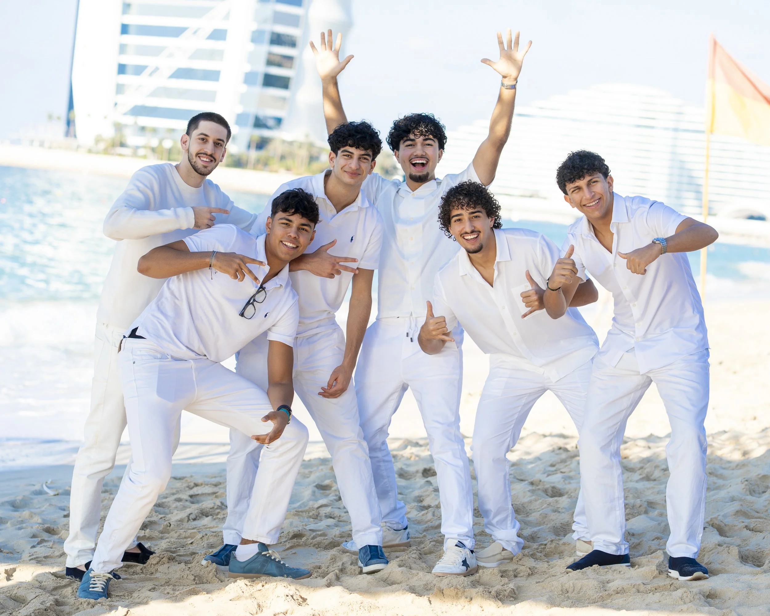 Six young men in white clothes posing and smiling on a beach with modern buildings and a blue sky in the background.