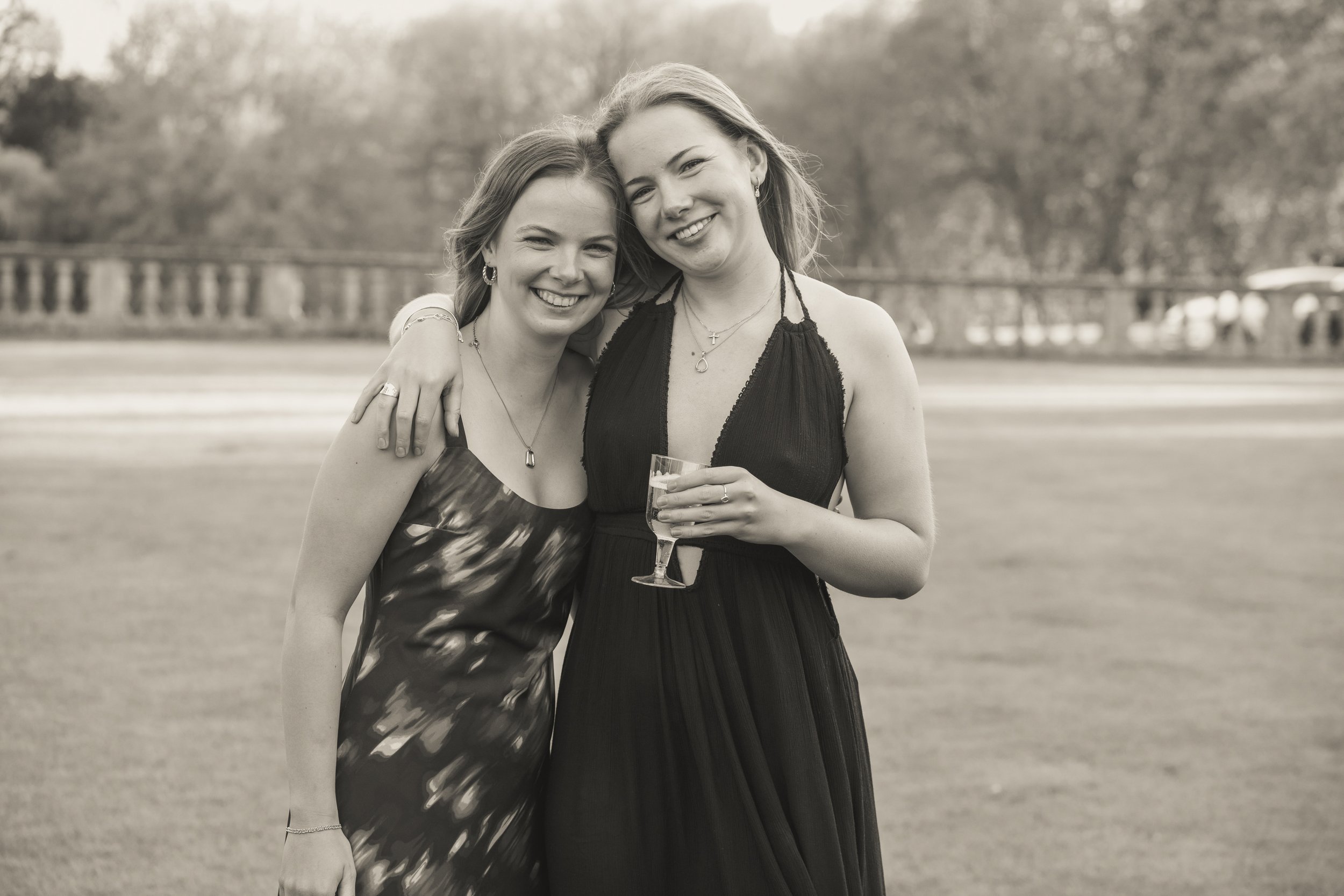 Two young women smiling, embracing, and holding a glass of champagne outdoors at an event, with a blurred background of trees and a fence.