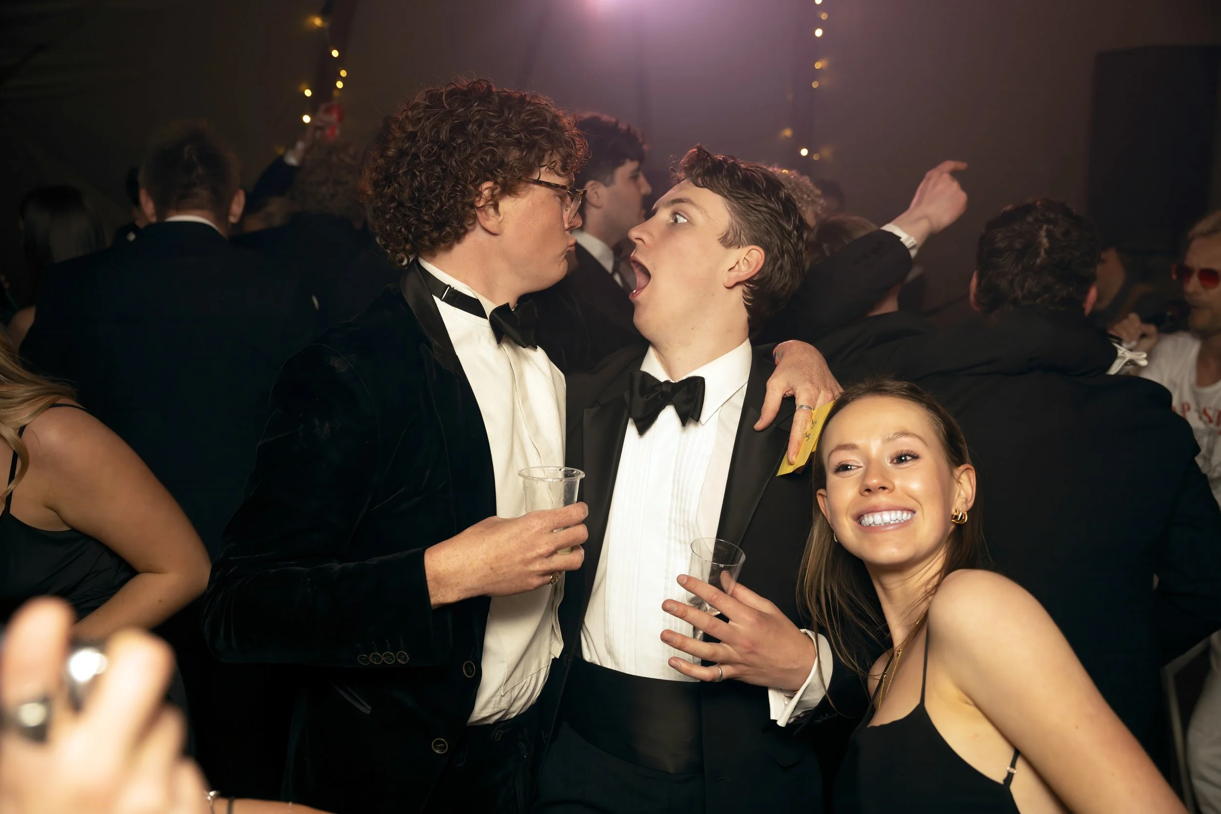 Three young adults dressed in formal tuxedos and evening wear at a party, with two men appearing to be having an animated conversation, and a smiling woman in a black dress in the foreground.