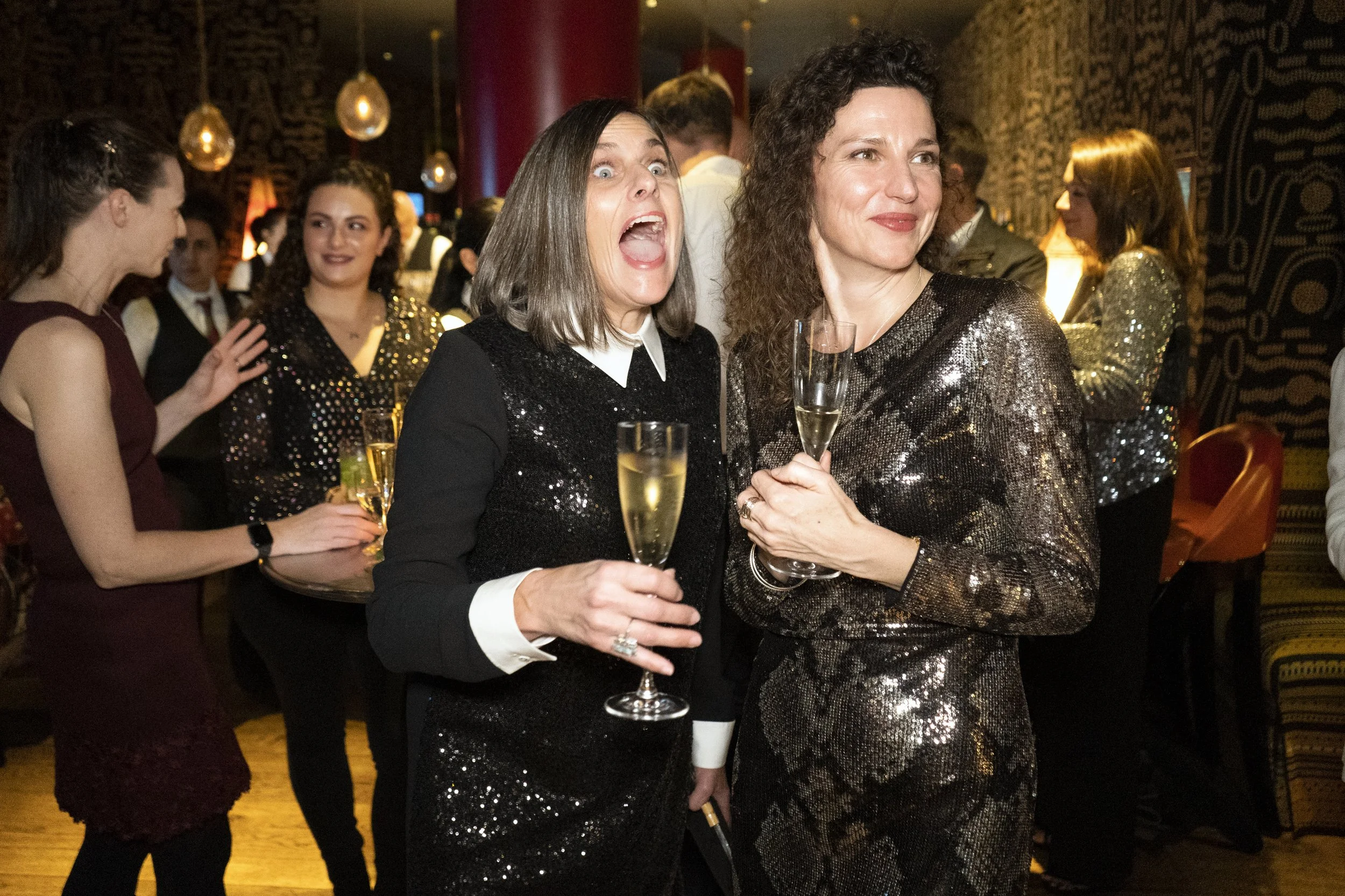 Two women holding champagne glasses at a party, one with a surprised expression and the other smiling, in a festive, dimly lit venue.