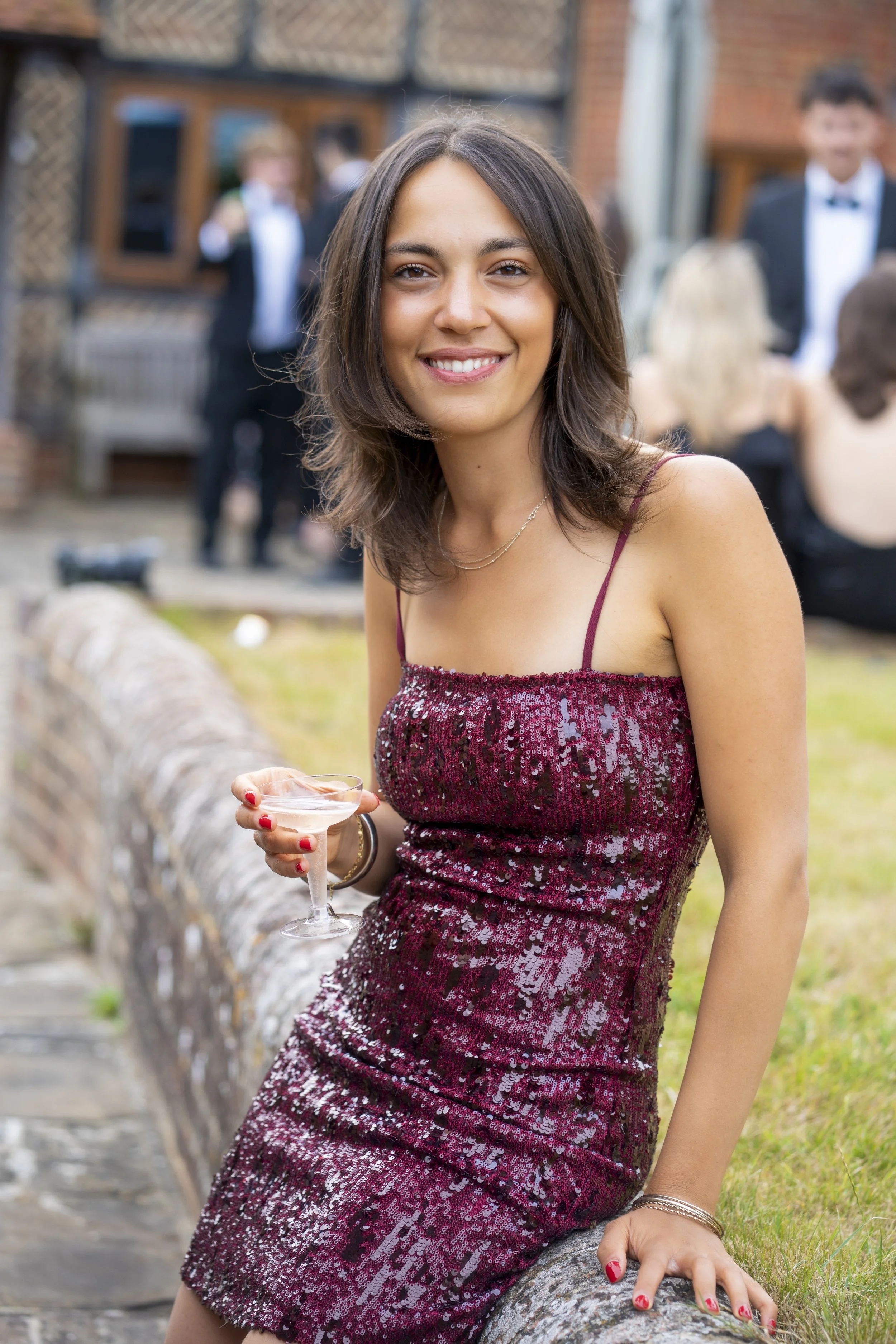 A woman in a sparkly maroon dress holding a cocktail at an outdoor party, with people dressed in formal attire in the background.