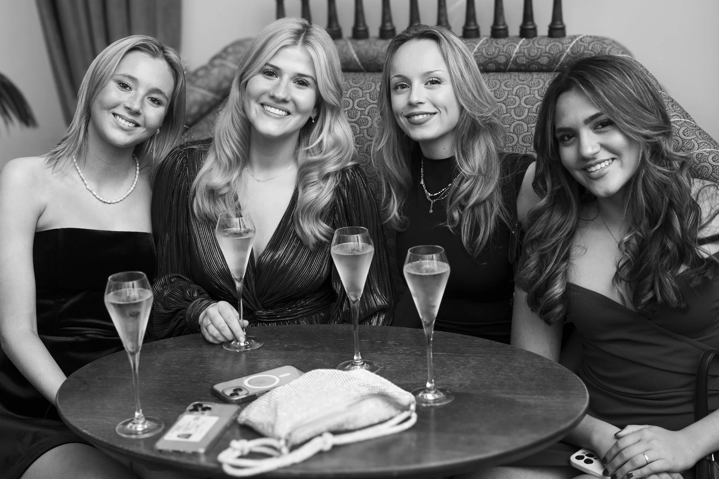 Four women sitting around a table with four glasses of champagne, smiling at the camera, in a celebratory setting.
