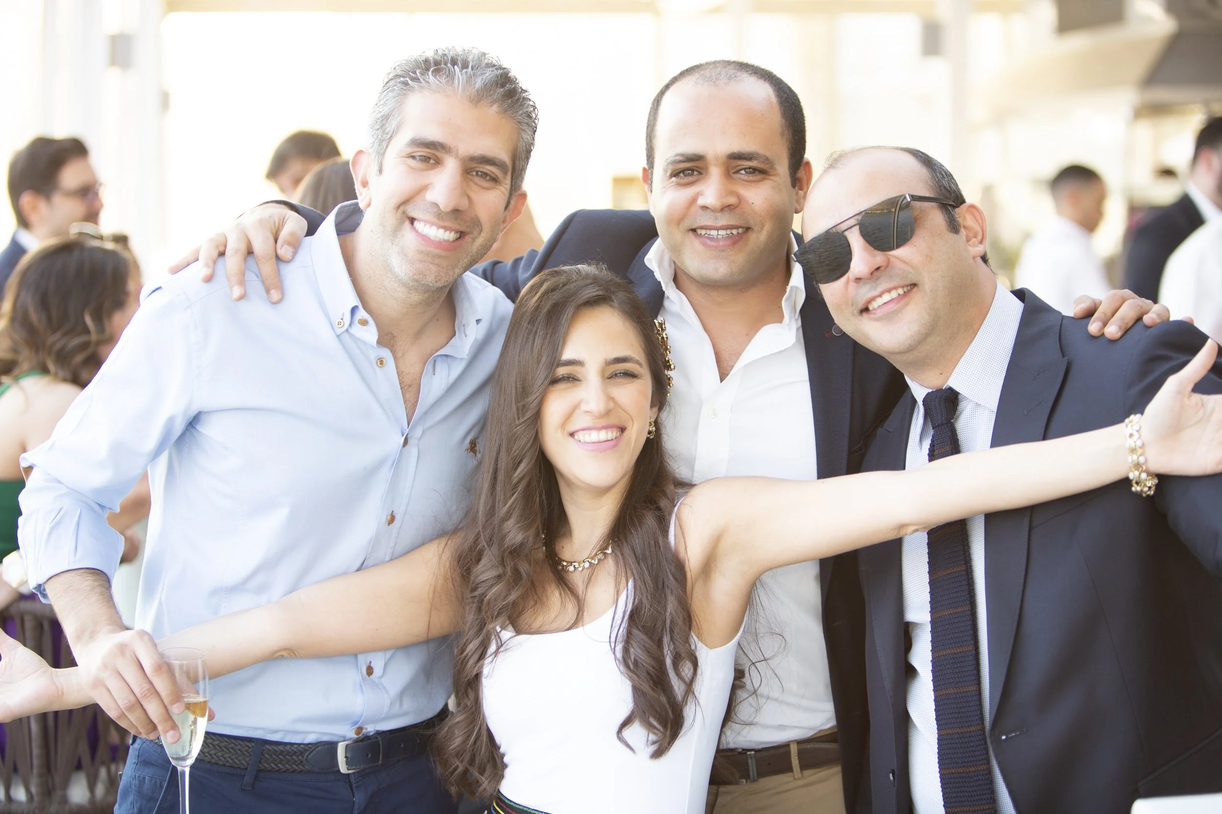 Group of five friends smiling, celebrating outdoors at a social event, with one woman reaching out and holding a glass of champagne.